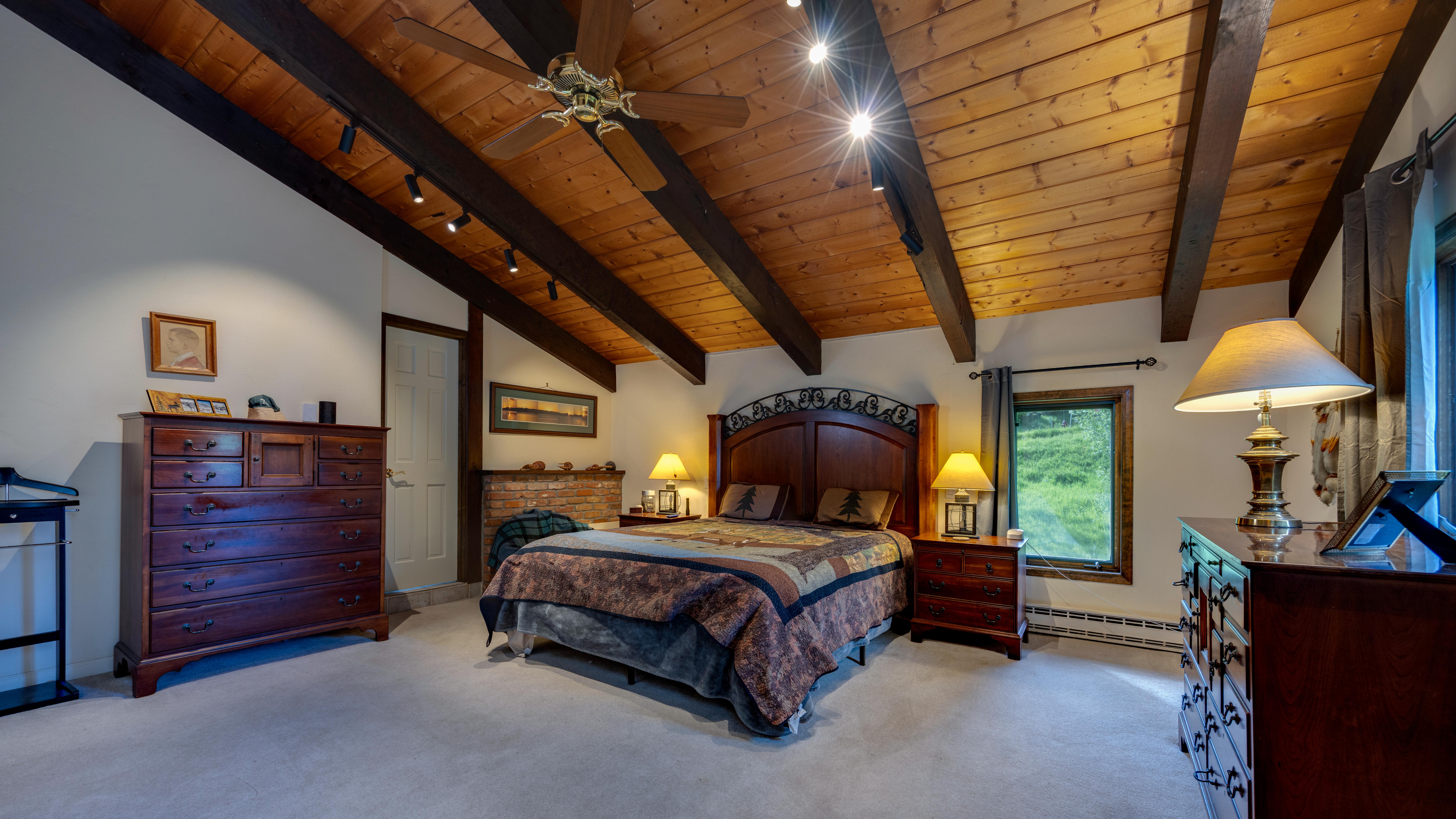 This spacious primary bedroom features a rustic design aesthetic with a vaulted wood-plank ceiling and exposed decorative beams. The room is centered around a rich, dark-wood headboard and a patterned quilt, complemented by dual nightstands with matching lamps and a large dresser providing ample storage. A window with forest views and warm recessed lighting complete the inviting and cozy atmosphere of this mountain-style space.
