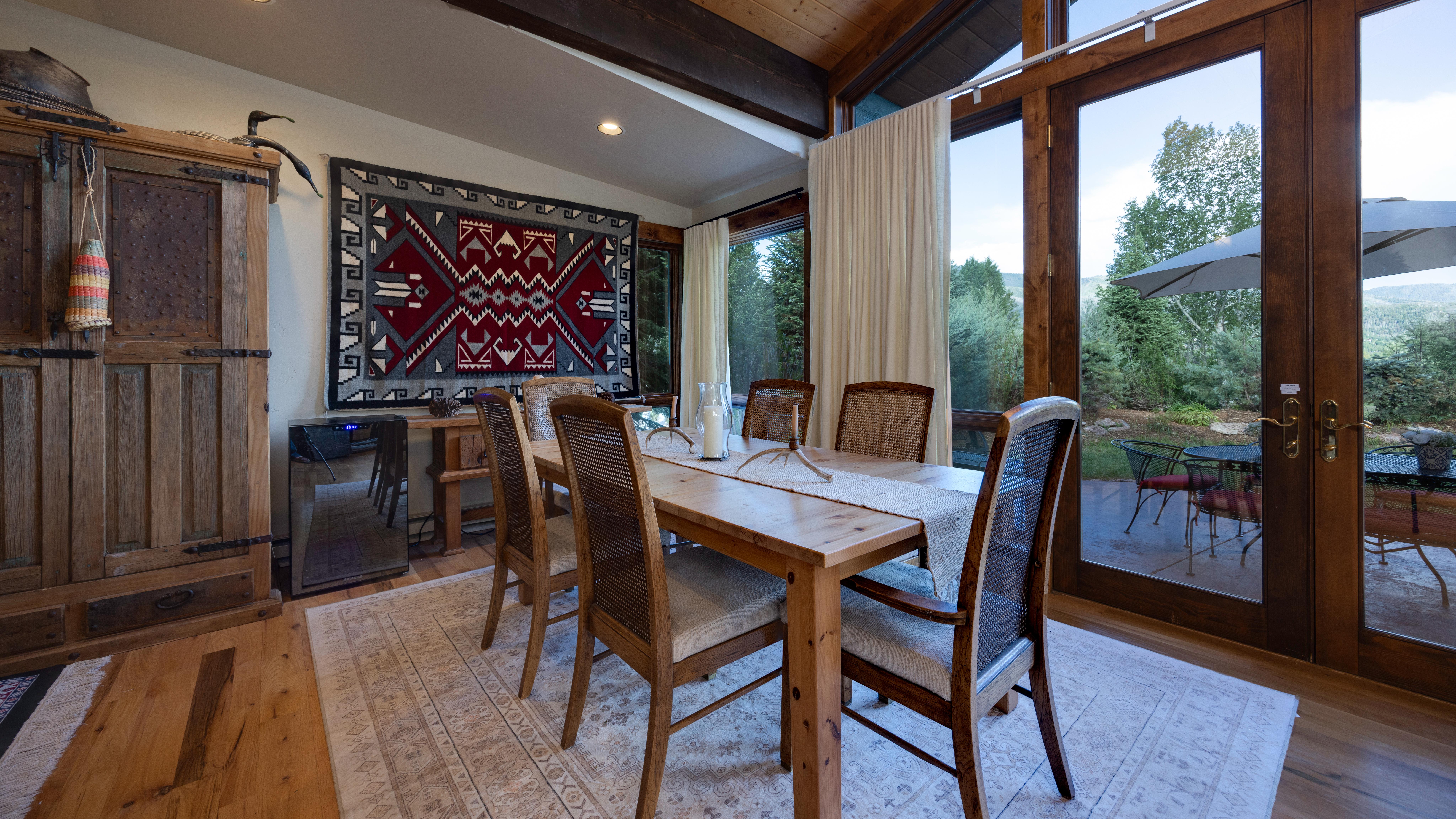 This warm and inviting dining room features a rustic wooden table with six cane-backed chairs set upon a neutral patterned area rug. A large, vibrant Native American-inspired textile hangs on the wall, adding a focal point, while floor-to-ceiling windows provide abundant natural light and seamless access to an outdoor patio. The space combines organic textures with traditional elements to create a cozy, sophisticated atmosphere.