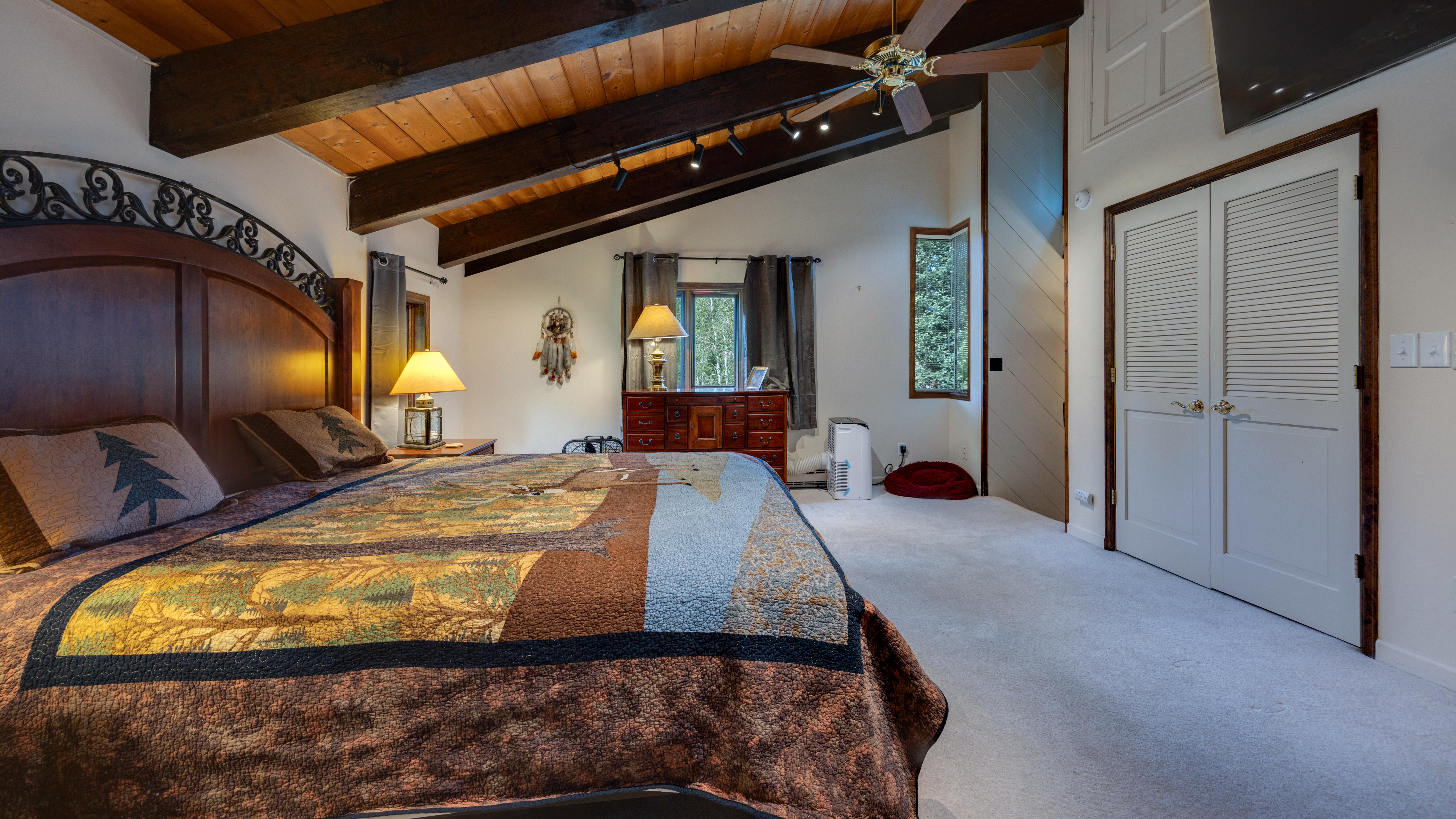 This spacious primary bedroom features a rustic-themed quilt and an ornate wrought-iron bed frame, set against a stunning vaulted wood-paneled ceiling with exposed beams. The room is warmly lit by two table lamps and a ceiling fan, offering a comfortable and cozy atmosphere. Notable features include a wooden dresser, a window with natural views, and white louvered closet doors, creating a perfect blend of cabin-style charm and functional living space.