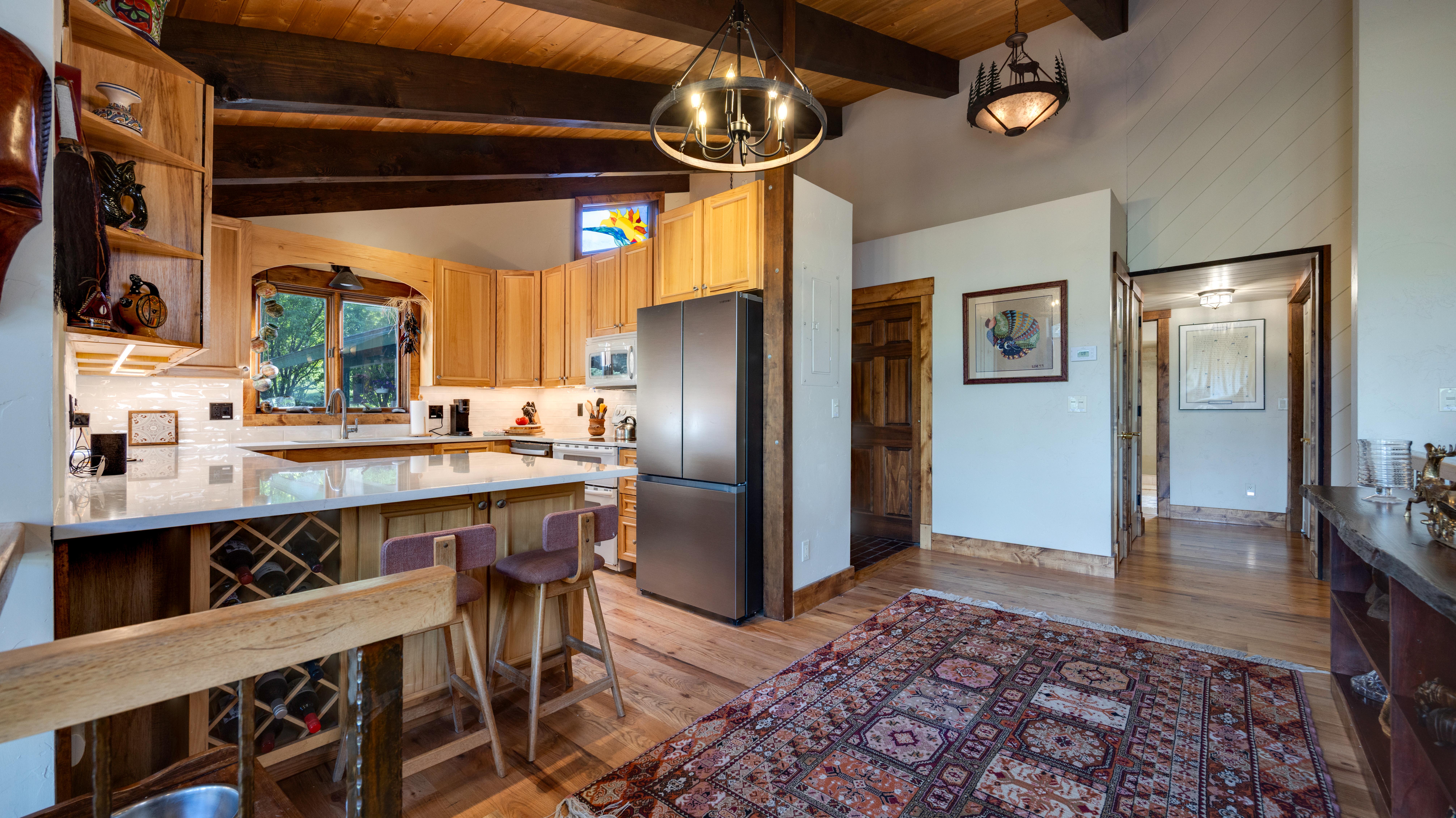 This rustic, custom wooden kitchen showcases vaulted ceilings with exposed beams, light wood cabinetry, and a spacious peninsula with built-in wine storage. The warm space is accented by unique stained glass detailing, professional-style kitchen lighting, and a patterned area rug that transitions into the adjacent hallway. Its natural material palette creates a cozy and inviting atmosphere typical of a high-end mountain or craftsman-style home.