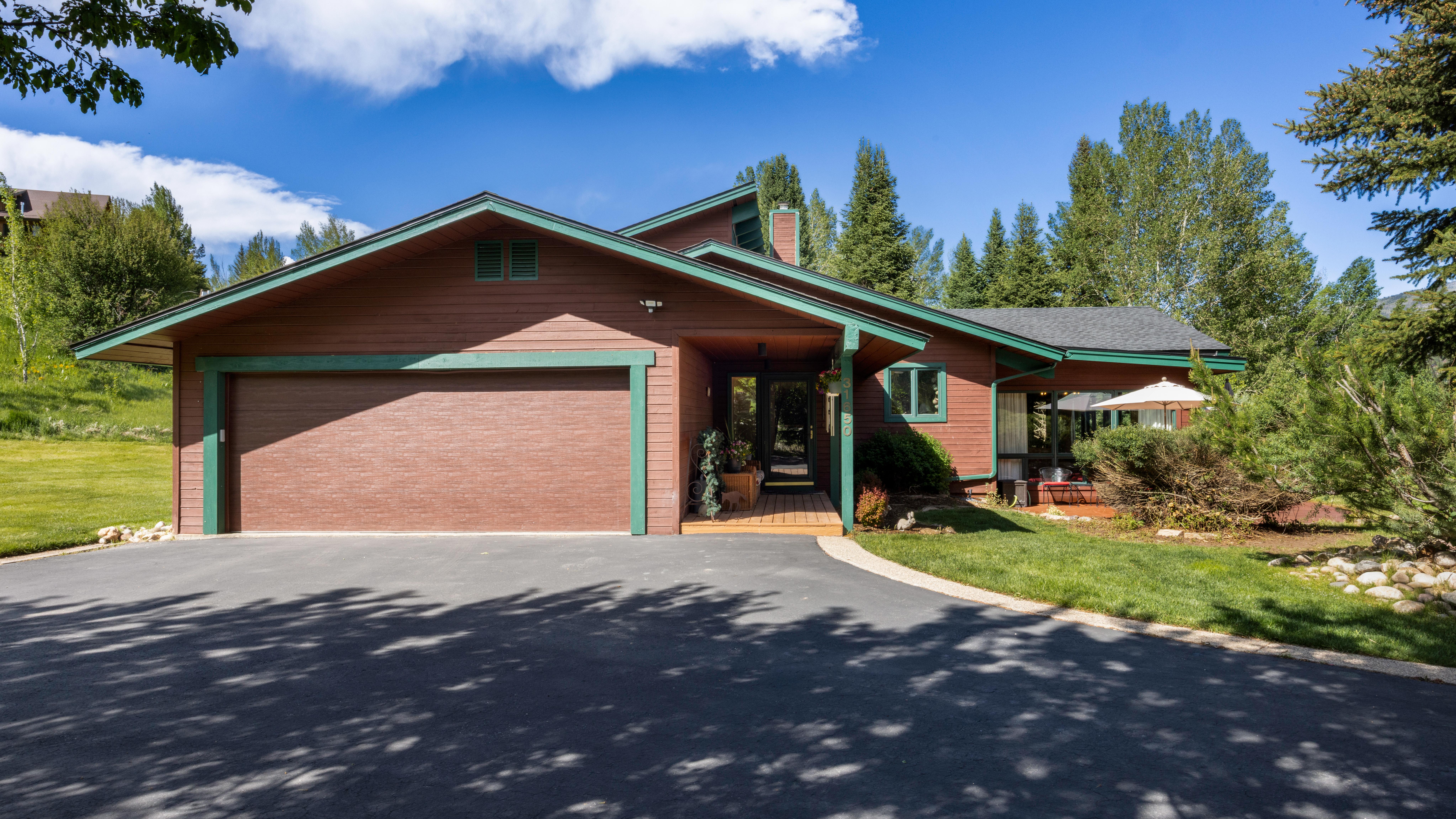 This charming, multi-level mountain home features warm brown wood siding accented by green trim and a spacious attached two-car garage. The front entrance is welcoming, leading to a recessed porch area set against a backdrop of mature pine trees and lush greenery. A paved driveway leads up to the home, creating a peaceful, established curb appeal in a serene natural setting.