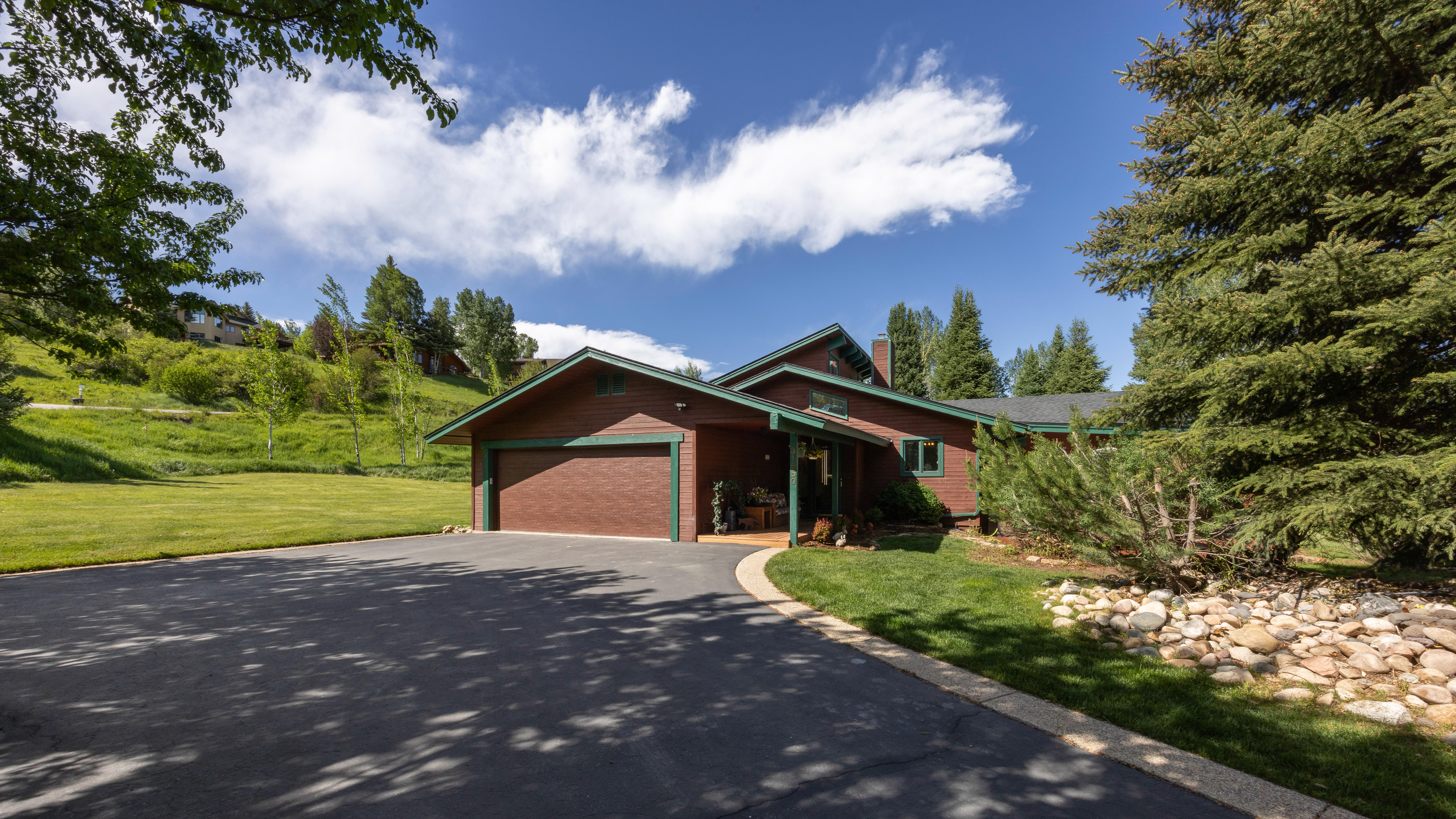This charming alpine-style home features rustic stained wood siding, green trim, and a prominent front-facing garage. The property is set against a lush, grassy hillside under a clear blue sky, accented by mature trees and a neatly landscaped rock garden. The paved driveway leads up to a recessed entryway, creating a warm and inviting look that blends seamlessly with the natural mountain environment.