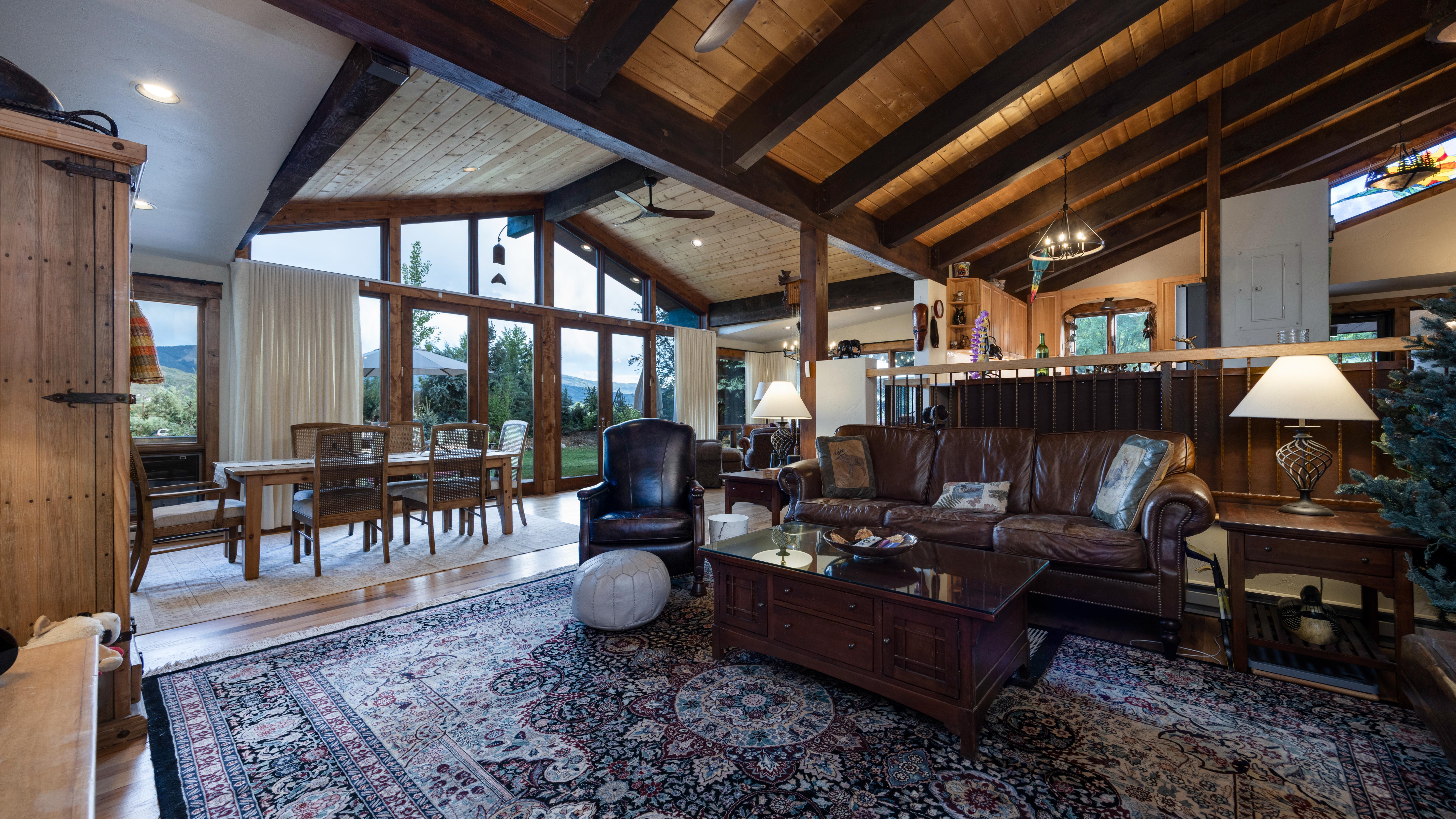 This spacious, open-concept living and dining area features vaulted, exposed-beam ceilings and extensive floor-to-ceiling windows that offer ample natural light. The room is anchored by a rich, dark leather sofa, a traditional Persian-style area rug, and a solid wood coffee table, creating a warm, mountain-lodge aesthetic. The seamless integration between the comfortable seating area and the adjacent dining space makes this home ideal for entertaining while showcasing architectural character.
