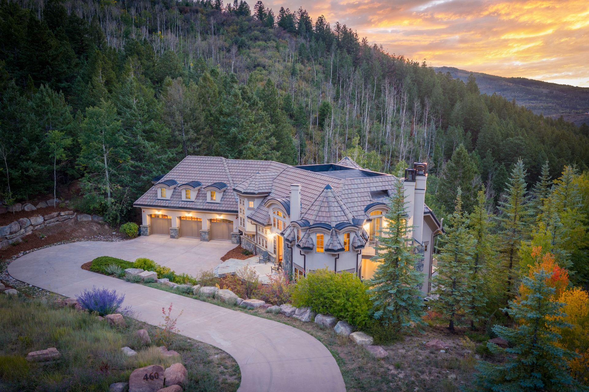 This is an aerial view of a luxurious home nestled in a mountain setting. The property features a long driveway, a three-car garage, and meticulously landscaped grounds. The architectural design includes a combination of stone and siding, with a roof that adds character, suggesting a high-end, private residential property.