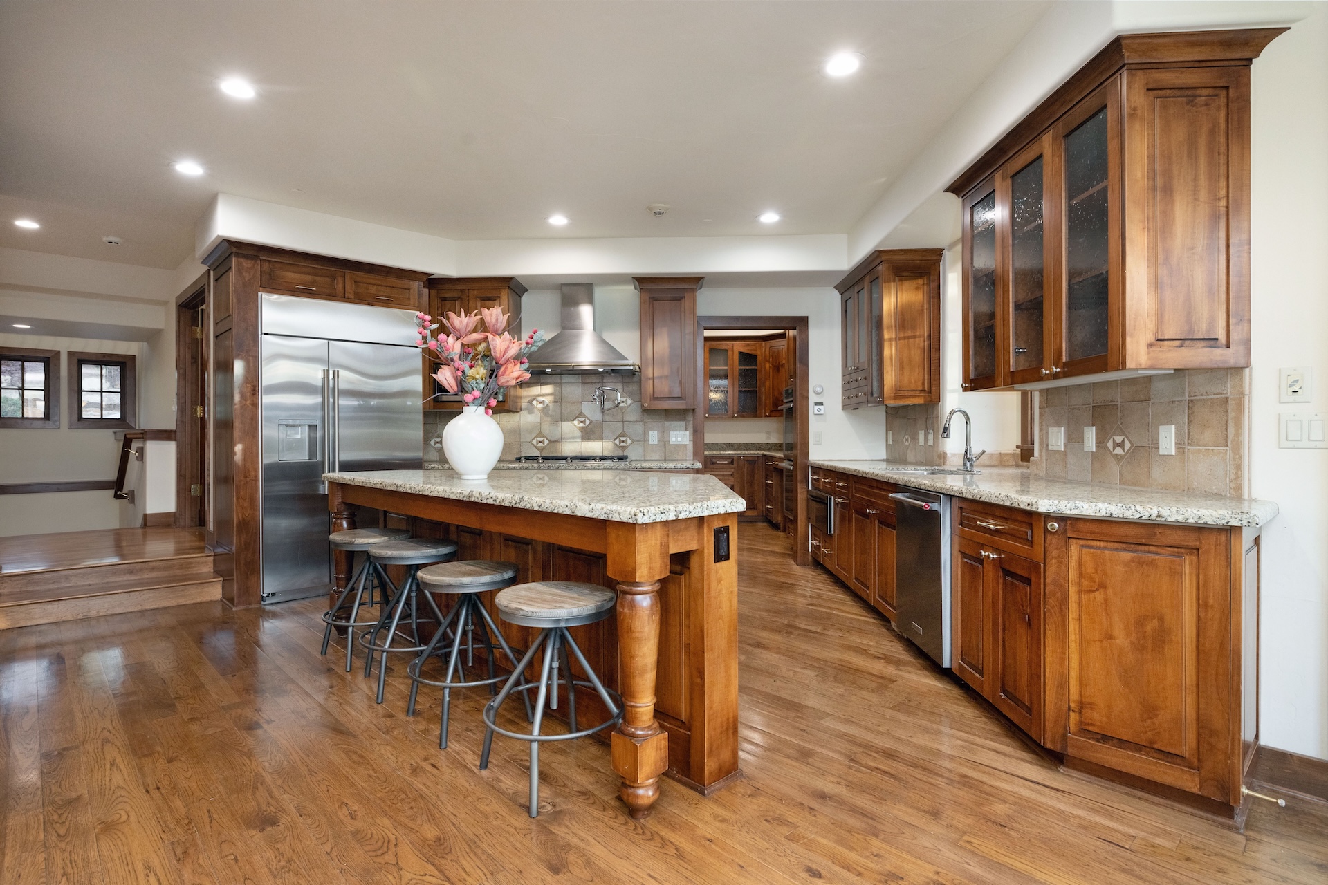 This is a well-maintained kitchen featuring wooden cabinetry, stainless steel appliances, and granite countertops. A large island offers seating, and the warm wood floors create a cozy atmosphere. The kitchen boasts ample storage and workspace, making it ideal for cooking and entertaining.