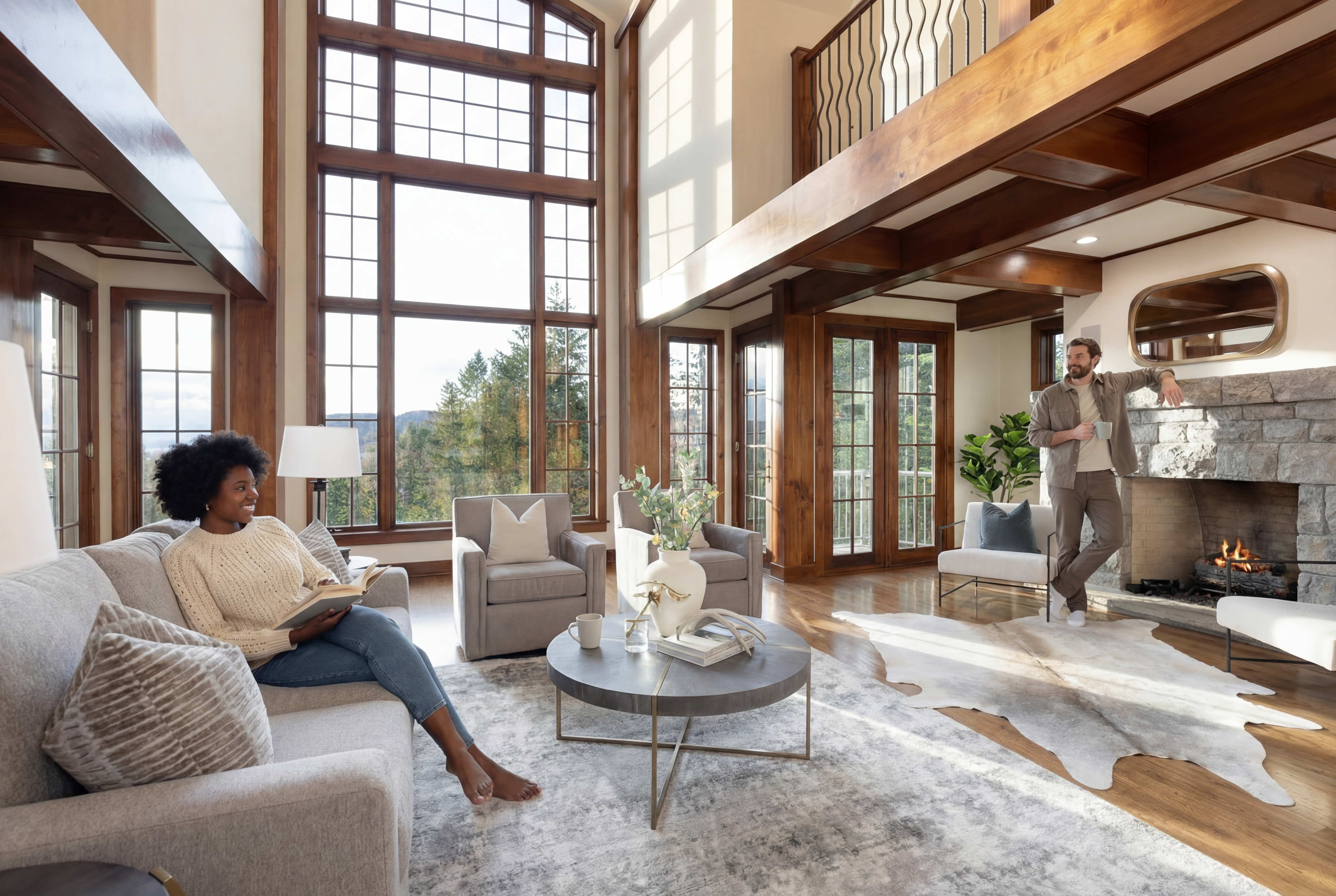 This is a bright and open living room with a large window offering scenic outside views and a stone fireplace. The room features comfortable seating, including a sofa and armchairs, arranged around a round coffee table on a patterned area rug. The architecture includes wooden beams and a loft area above, lending a warm and luxurious feel to the space.
