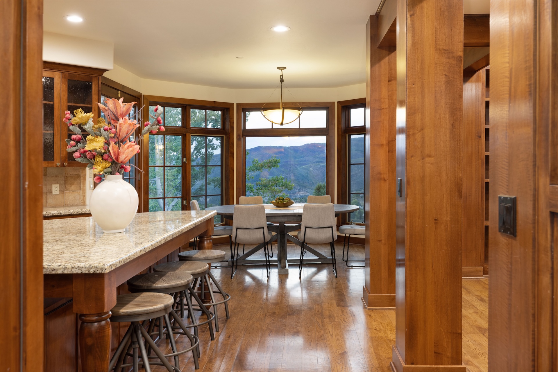 This inviting kitchen showcases warm wood tones and ample natural light. A granite-topped island with seating provides a central gathering point. The adjacent dining area boasts panoramic window views, creating a seamless transition between cooking and dining spaces.