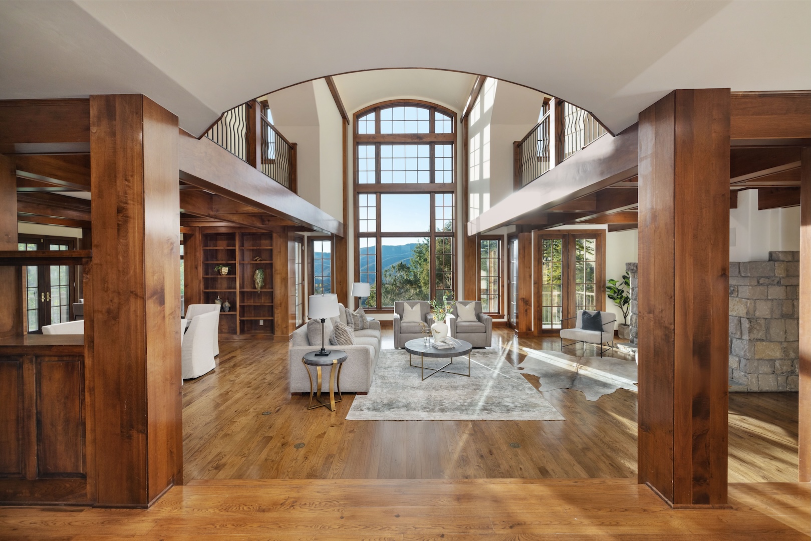 This expansive living room features a double-height ceiling with a large window offering views of the mountains. The room is furnished with a neutral-colored sofa, armchairs, and a circular coffee table, all arranged on a patterned rug. Wood flooring and ceiling beams add warmth and character to the space, creating a sophisticated and inviting atmosphere.
