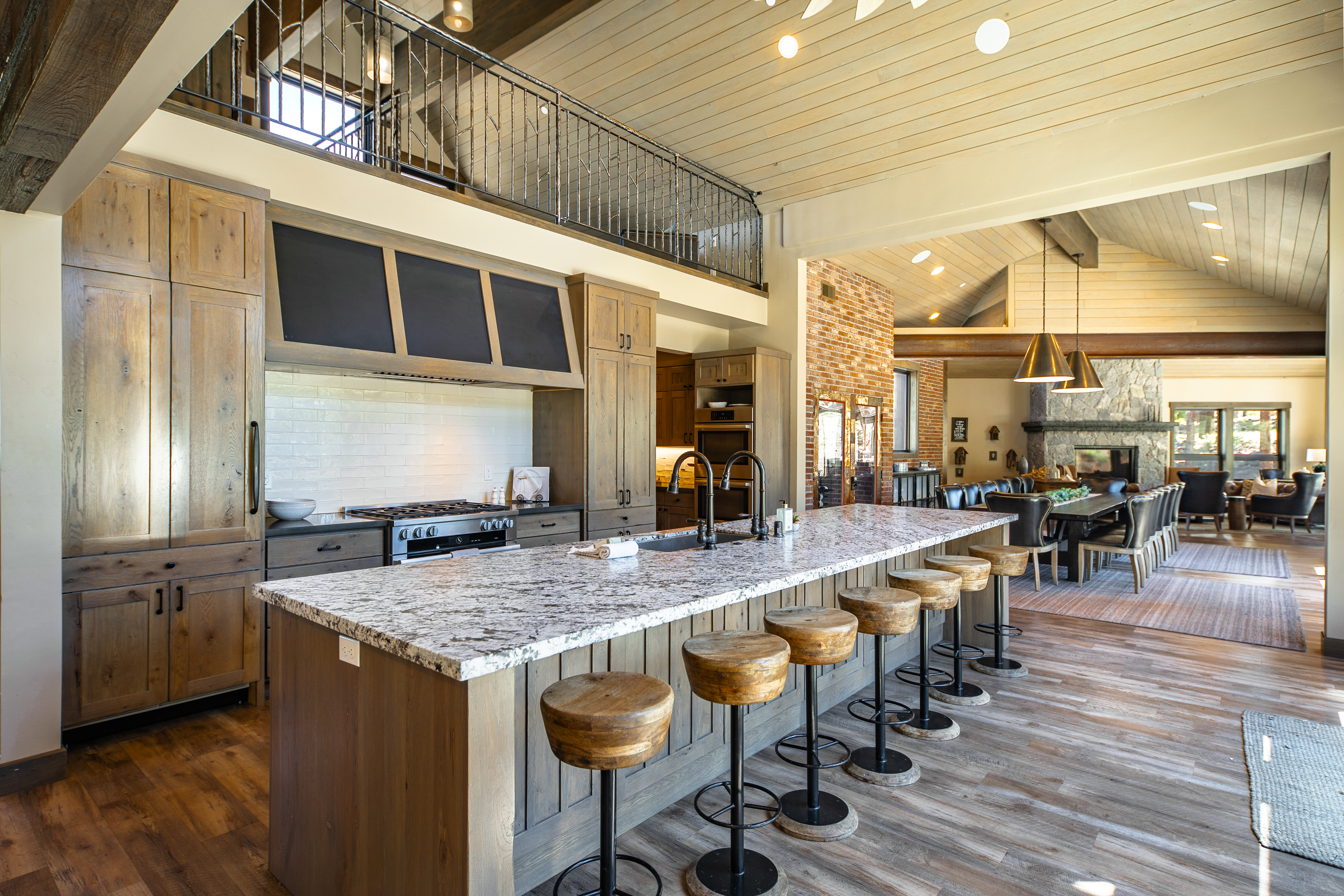 This expansive kitchen features custom wood cabinetry, a large center island with seating, granite countertops, and high-end appliances. The rustic yet elegant design includes hardwood flooring and extends into the dining and living areas, creating an open concept living space. A dramatic range hood and second-floor mezzanine add unique architectural interest.
