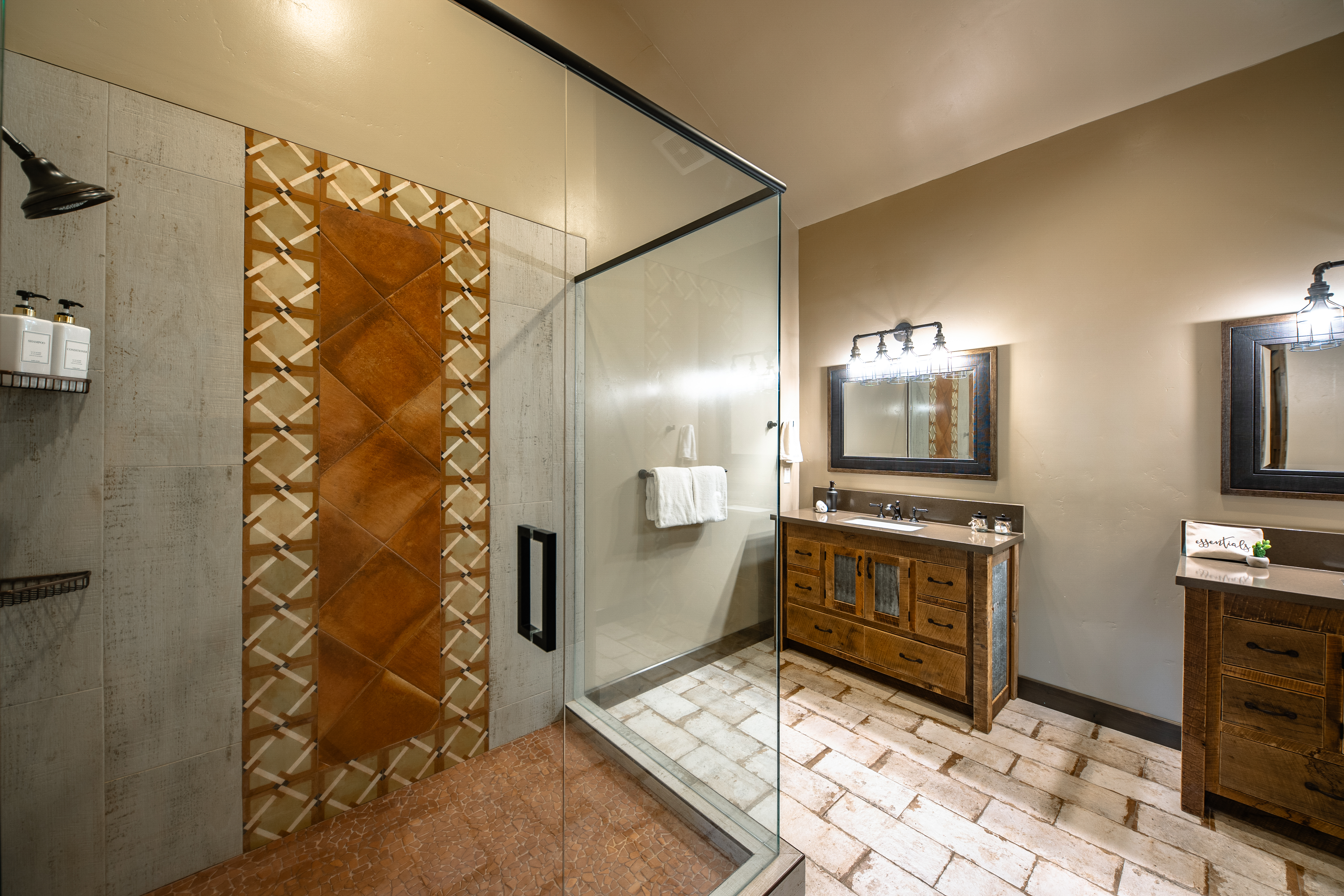 This is a well-appointed primary bathroom featuring rustic-style cabinetry with stone countertops and two framed mirrors illuminated by sconces. The shower is enclosed in glass and features designer tile work. Terracotta-colored flooring adds warmth and character to the space.