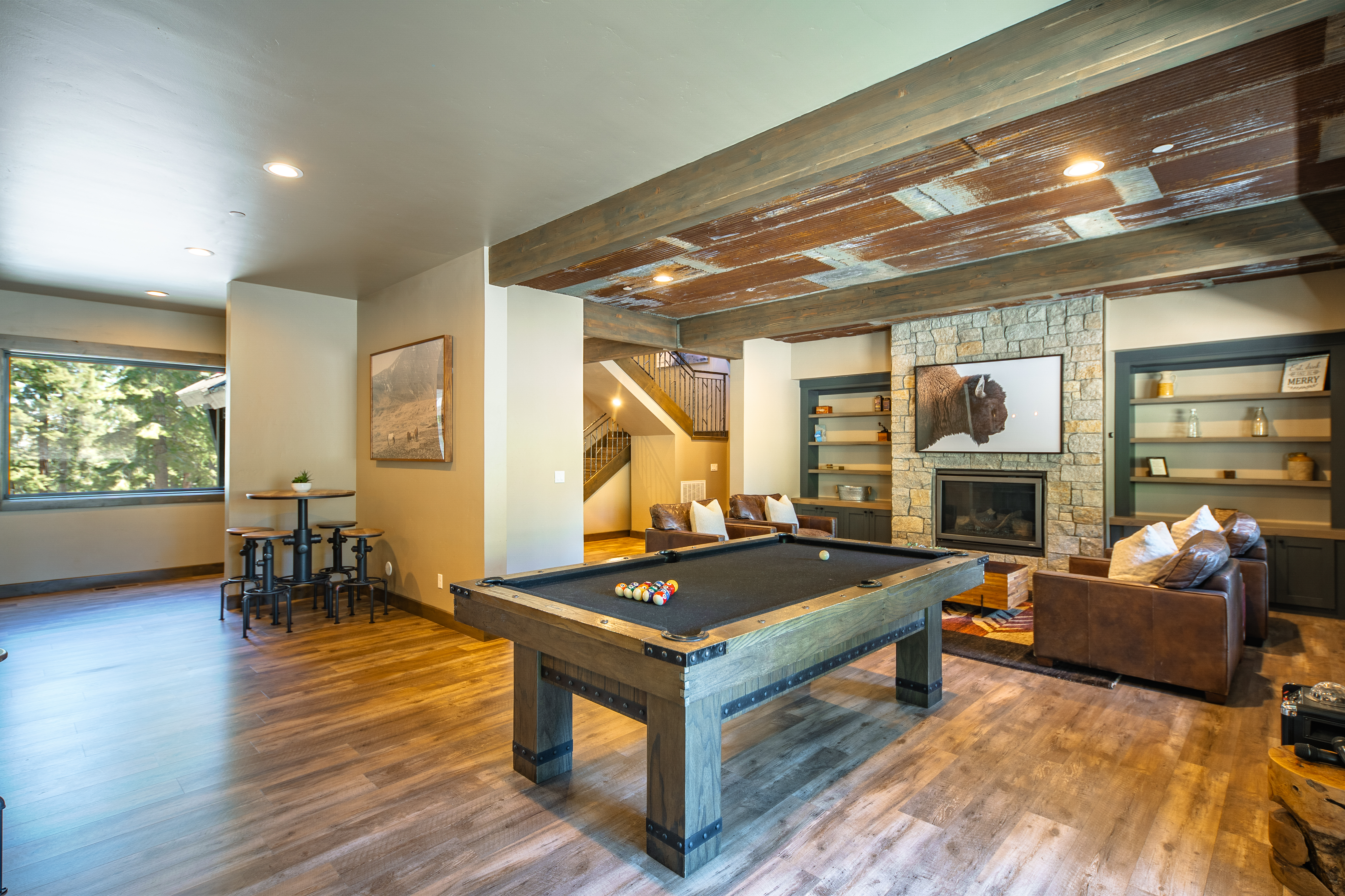 This is an inviting living room featuring a billiards table as a central feature. The room also includes comfortable leather seating arranged around a stone fireplace, and custom-built shelving. Wood flooring and a ceiling with wooden beams contribute to the rustic, yet sophisticated atmosphere of the space.