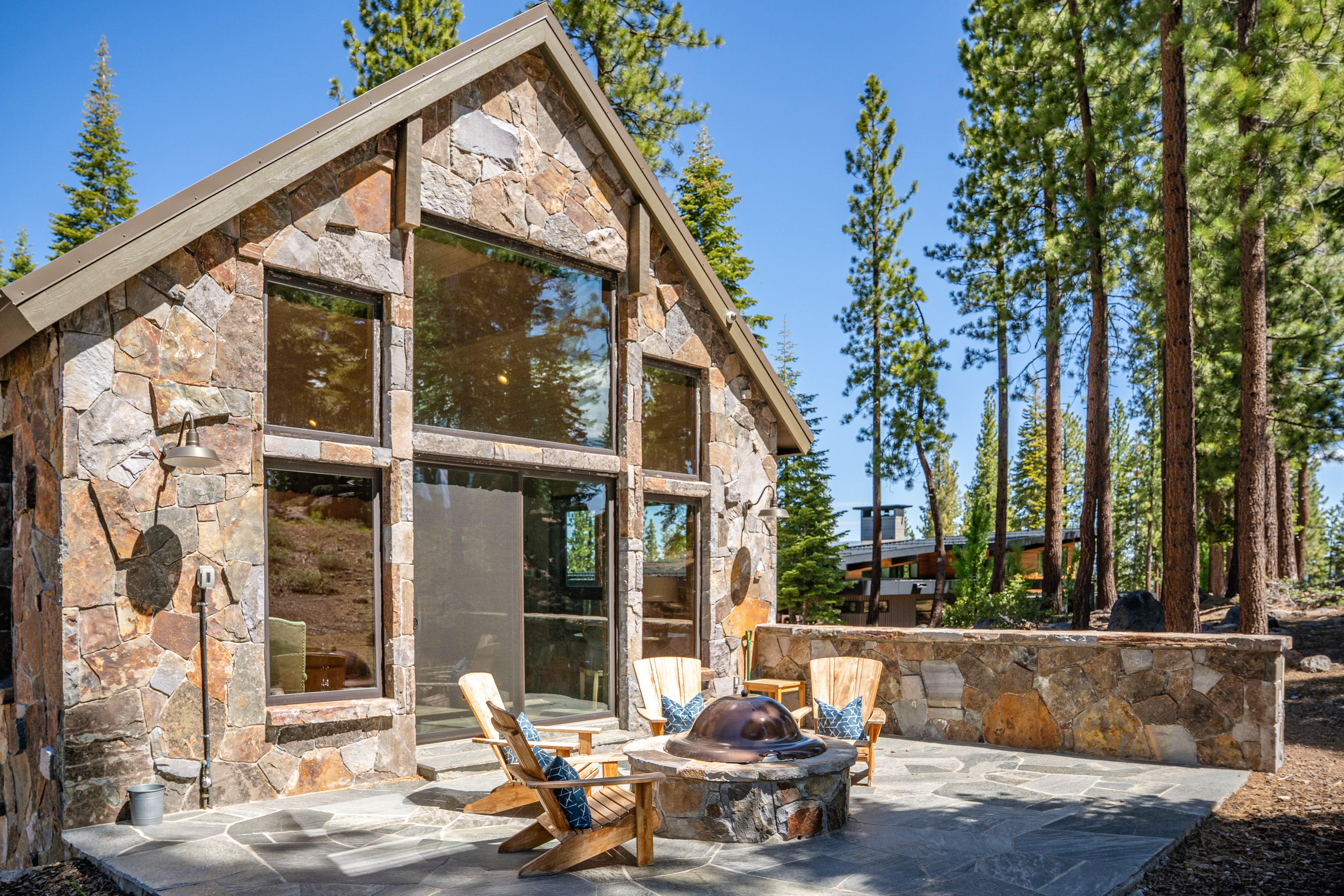 This image showcases a stylish rear patio area of a stone-clad home. A stone fire pit is surrounded by wooden Adirondack chairs, suggesting a cozy outdoor living space. Tall trees in the background provide privacy, enhancing the inviting atmosphere of this outdoor recreational area.