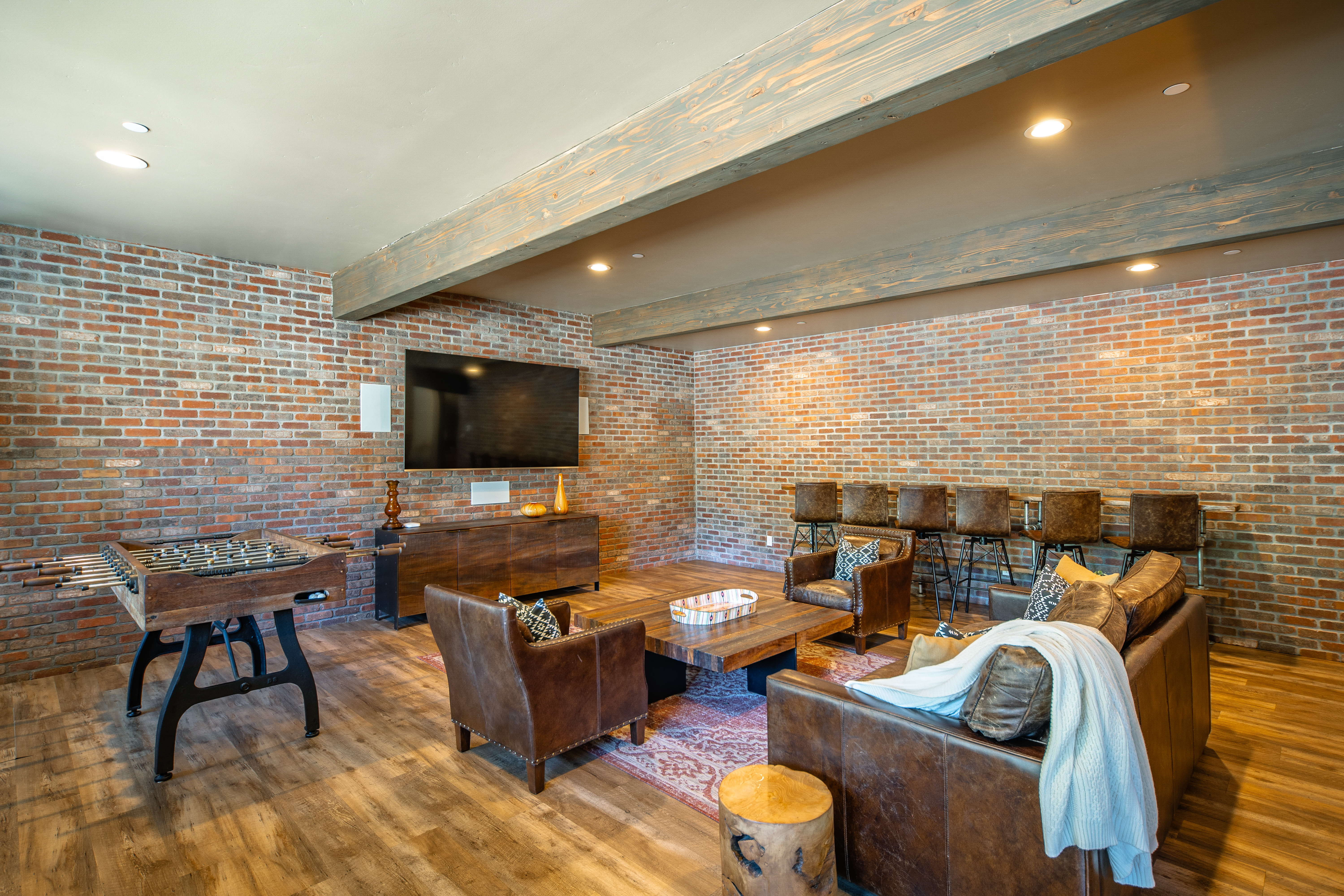This image showcases a living room featuring exposed brick walls, lending a rustic-industrial vibe. The space includes comfortable leather seating, a wooden coffee table, and a foosball table, creating a relaxed and entertaining atmosphere. Wooden beams accent the ceiling, adding to the room's character and appeal.
