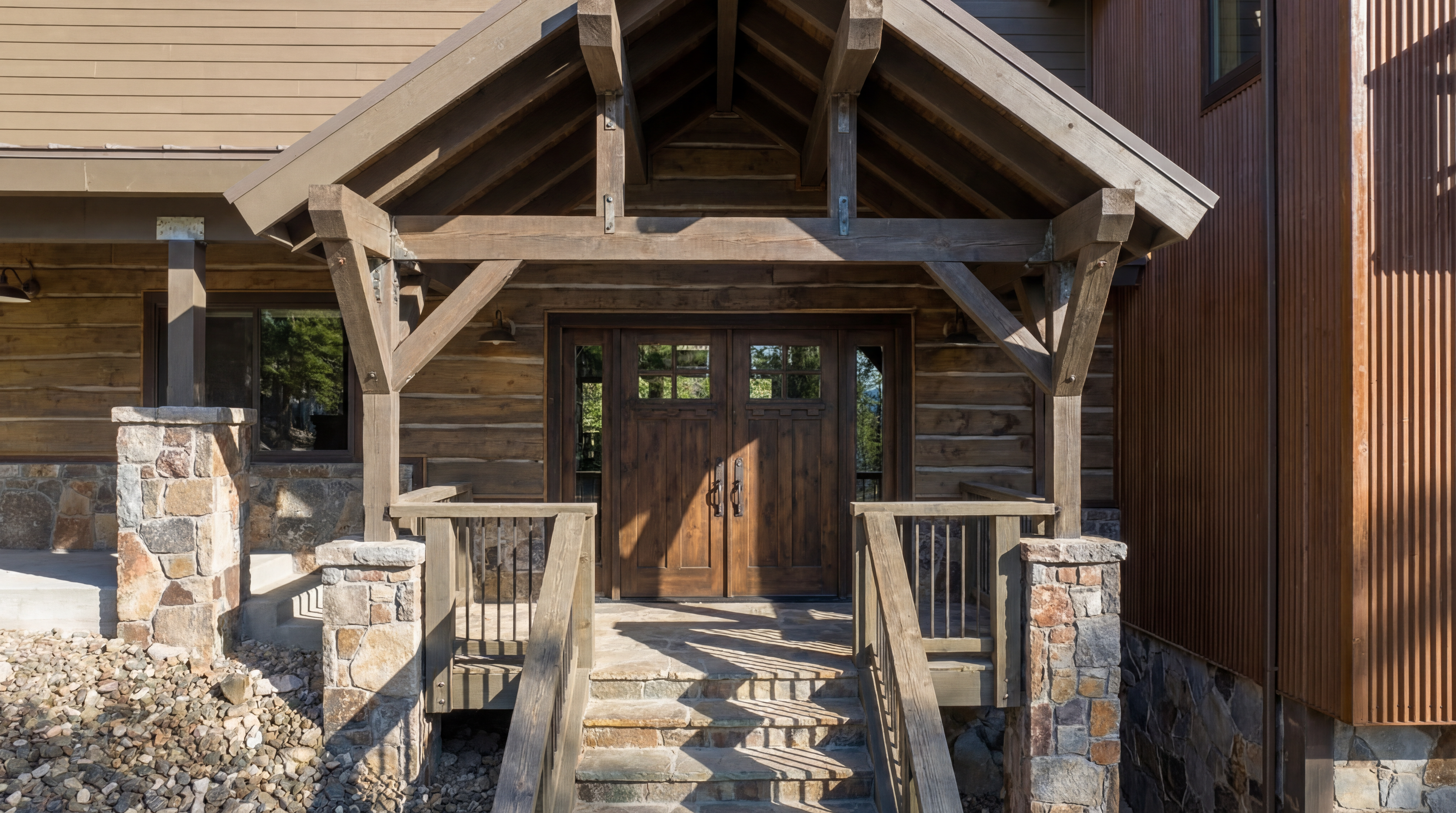 This image shows the entryway of a rustic-style home. The entrance features a wooden porch roof supported by stone pillars and wooden beams. The double doors are made of dark wood with small window panes, and stone steps lead up to the entrance, blending seamlessly with the landscape.