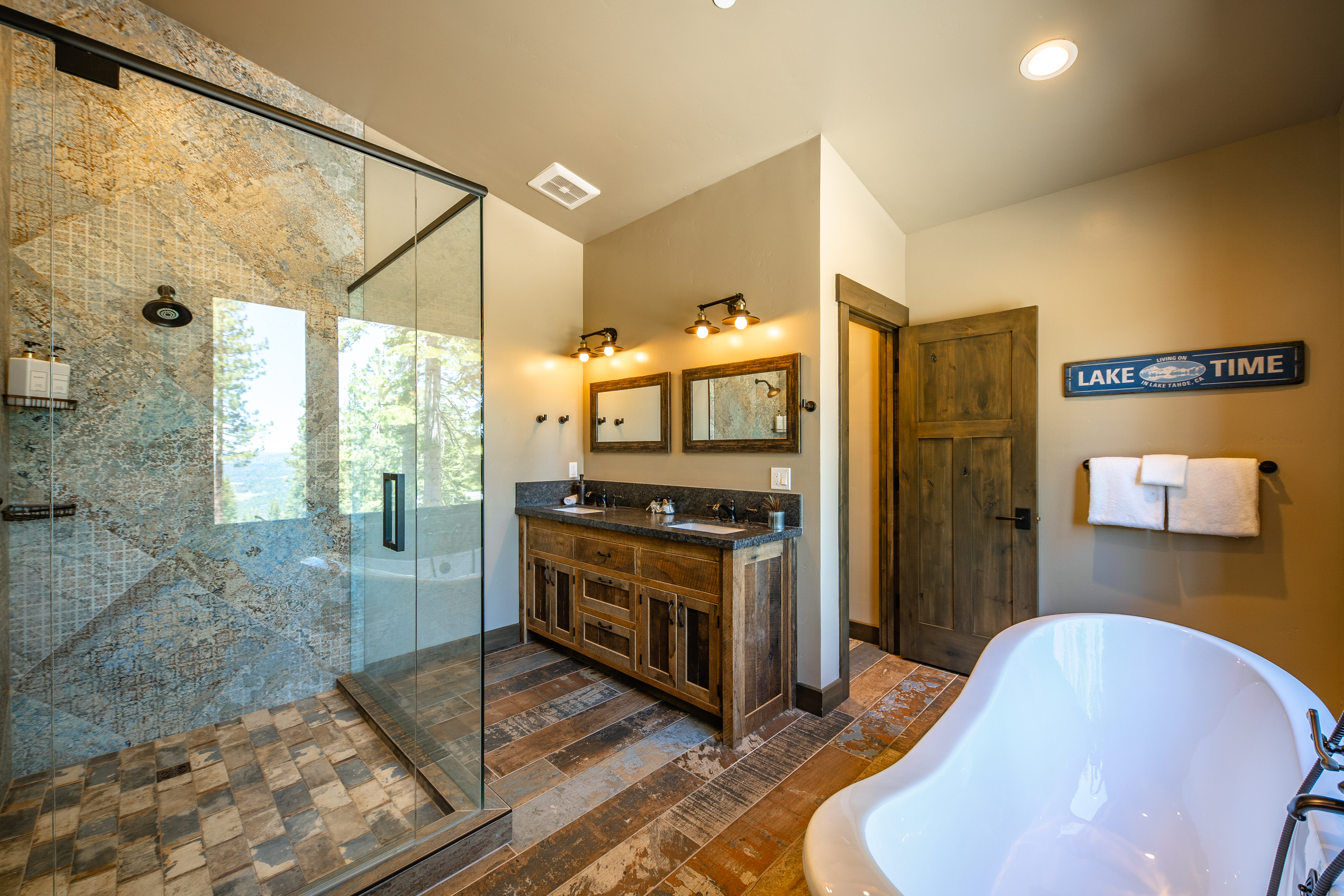 This is a bathroom showcasing a double vanity with a wood style cabinet, granite countertop and undermount sinks. A glass enclosed shower with uniquely patterned stone-style wall tiles sits beside the vanity. A stand-alone bathtub is partly visible in the lower right of the photo, with wooden floor tiles and a wall sign that reads "Lake Time" suggesting a rustic yet luxury style.