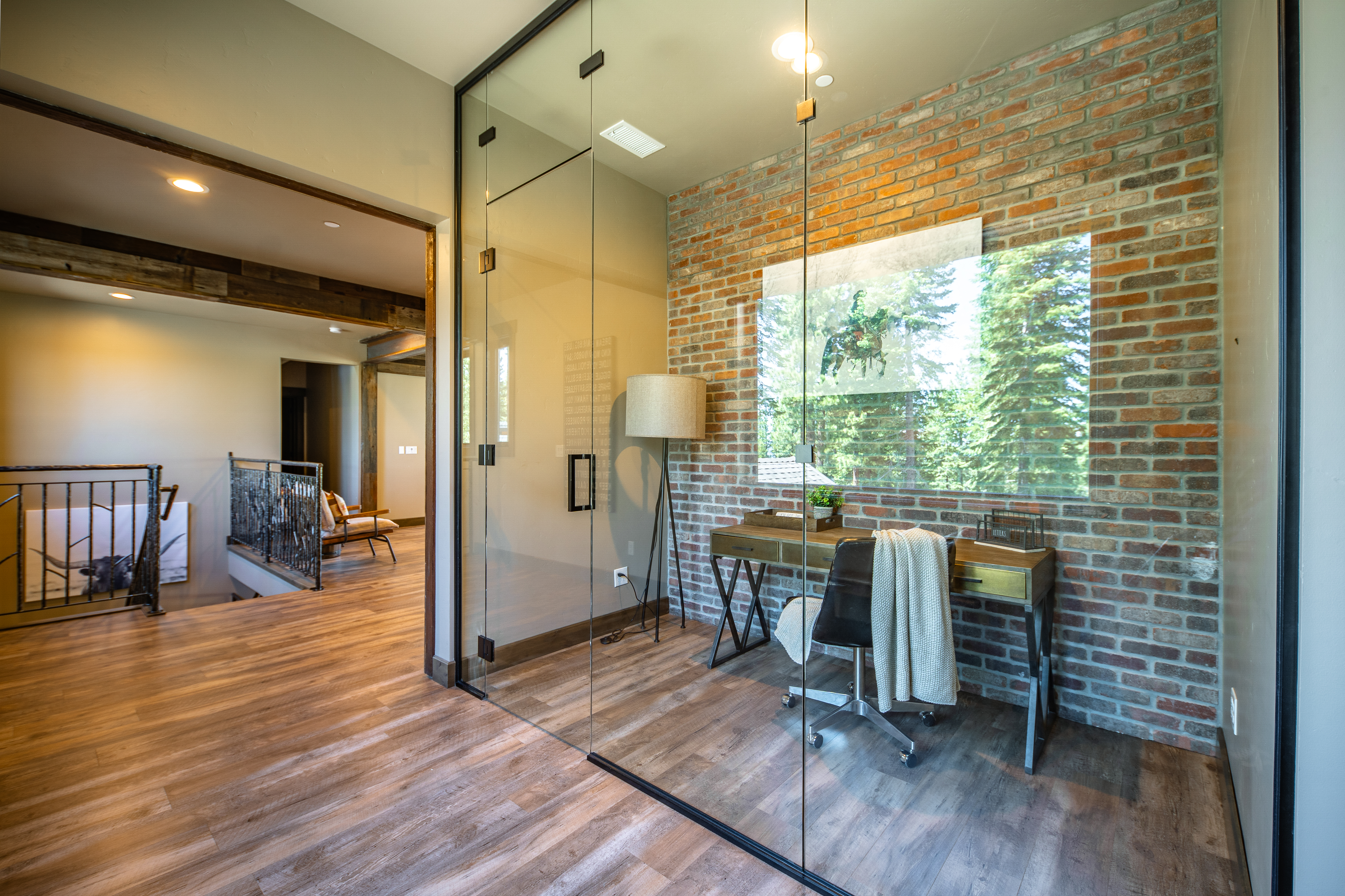 This is a modern office space with a brick accent wall and a glass partition, providing natural light and a sense of openness. The desk is neatly organized, and the room is furnished with a comfortable office chair and a floor lamp. The overall design is stylish and functional, making it an appealing workspace.