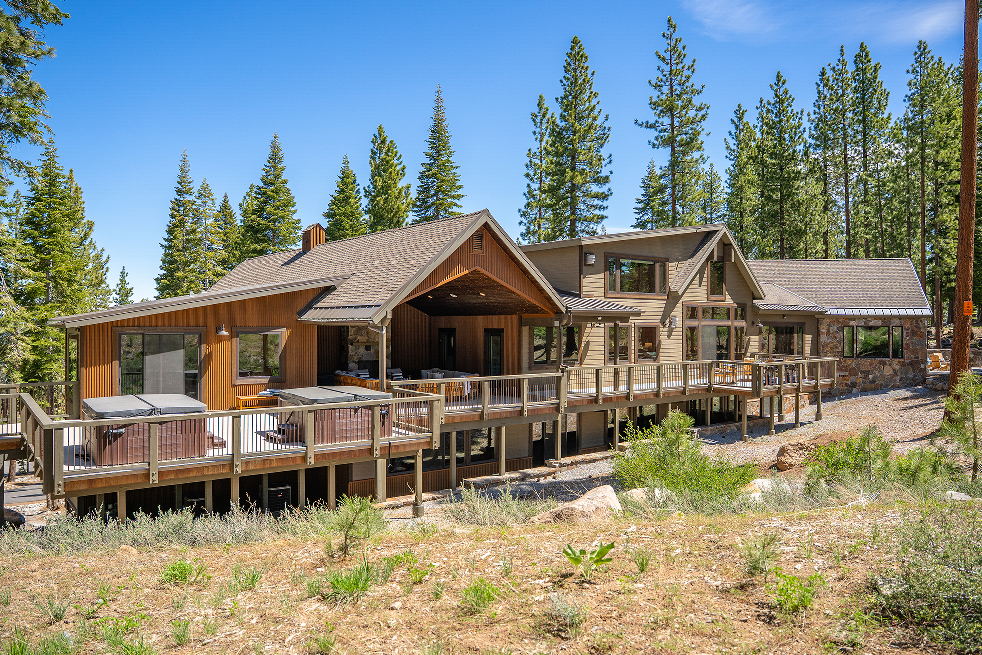 This image showcases the rear of a luxurious mountain home with an extensive deck featuring multiple hot tubs. The architectural details include wood siding, stone accents, and a gabled roofline, blending seamlessly with the surrounding forest. The overall impression is one of upscale mountain living with a focus on outdoor enjoyment and privacy.
