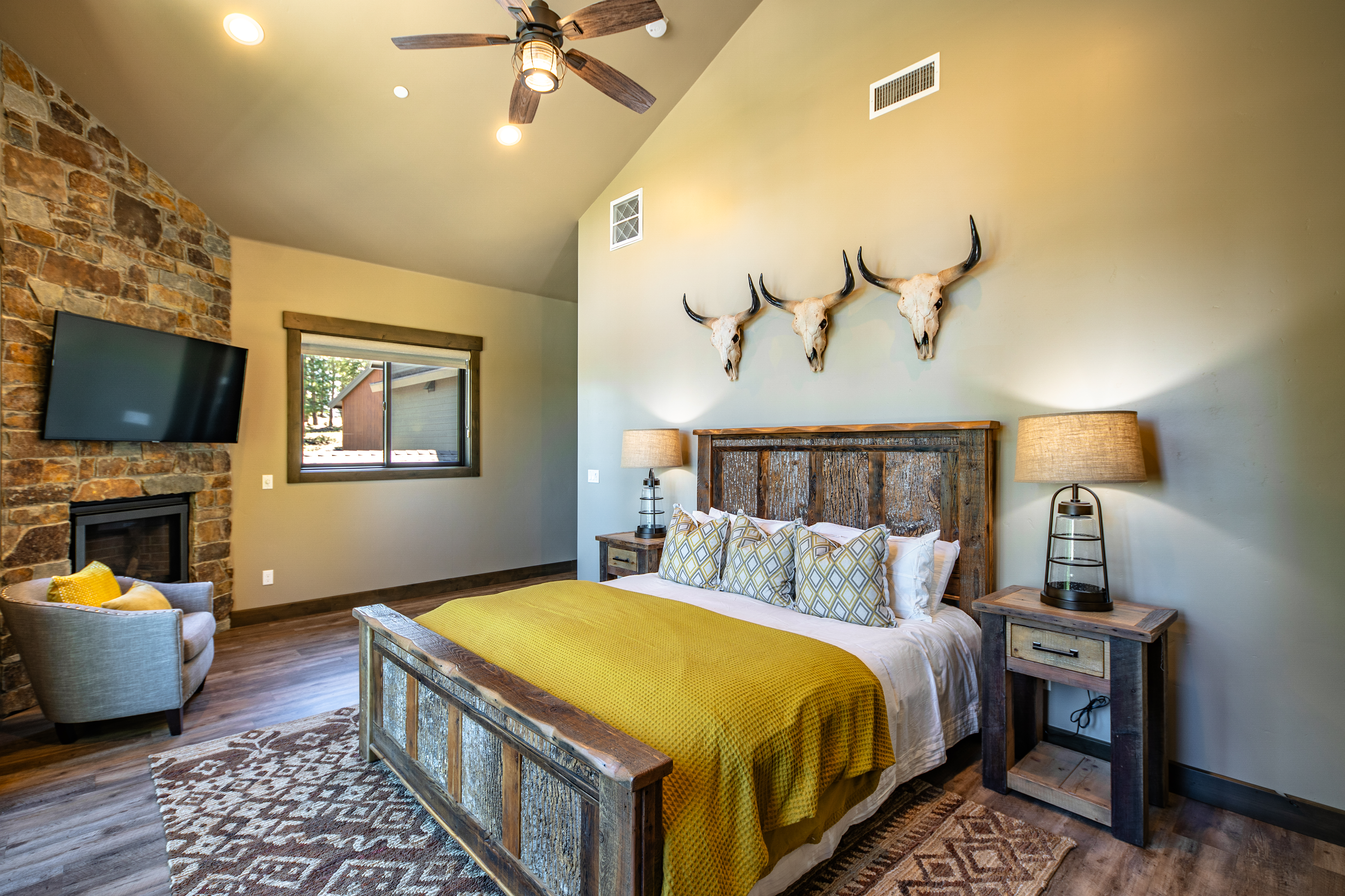 This is a primary bedroom featuring a rustic-style wooden bed with a yellow throw blanket. The room has a vaulted ceiling, a stone fireplace with a modern TV above, and three steer skulls mounted on the wall. The overall impression is cozy and stylish with a lodge-like aesthetic.