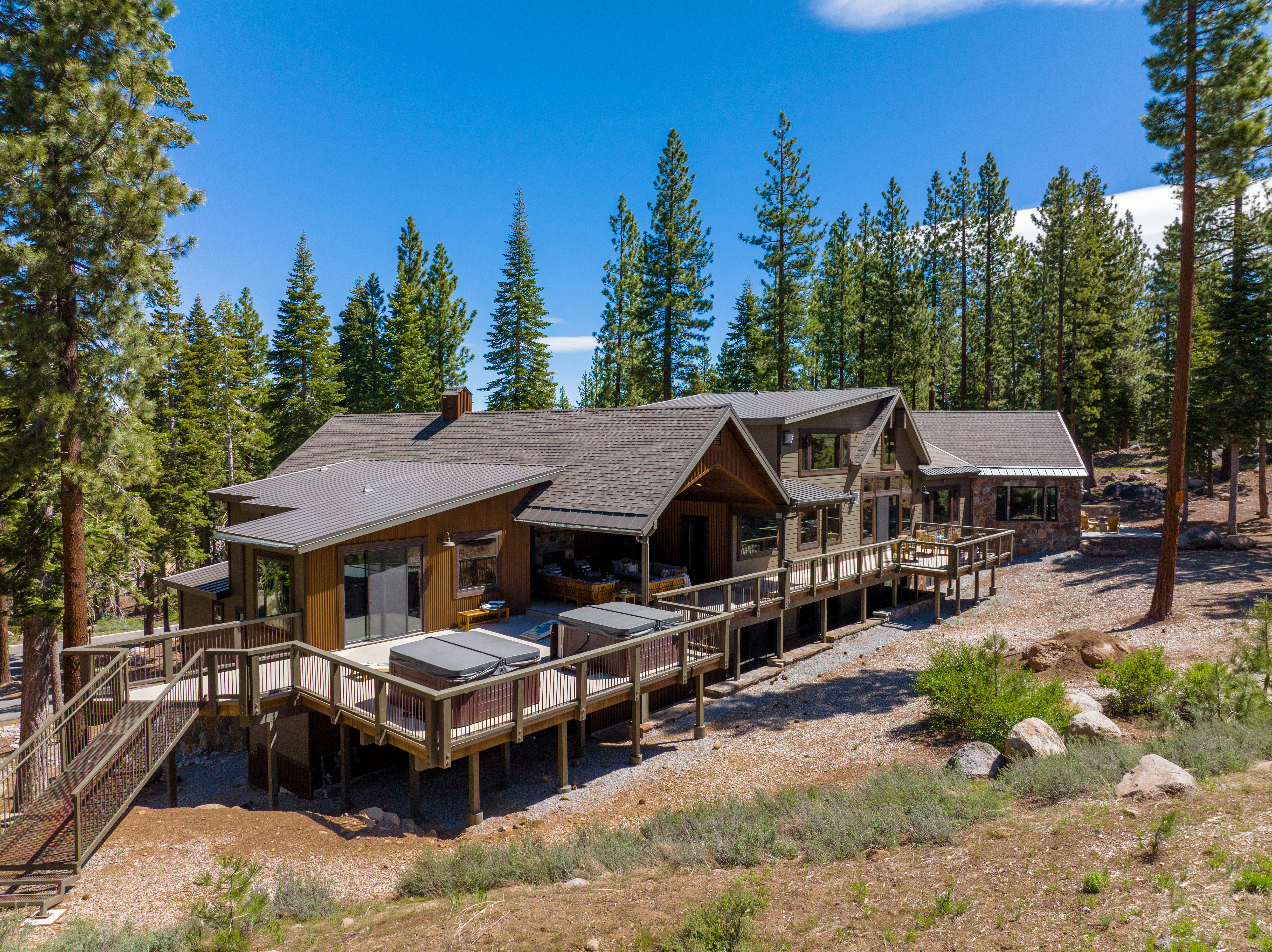 This image showcases the rear exterior of a beautifully designed home, featuring an expansive deck with multiple hot tubs. The house is surrounded by tall evergreen trees under a clear blue sky, offering a sense of privacy and tranquility. The decks and balconies suggest ample outdoor living space for relaxation and entertainment.