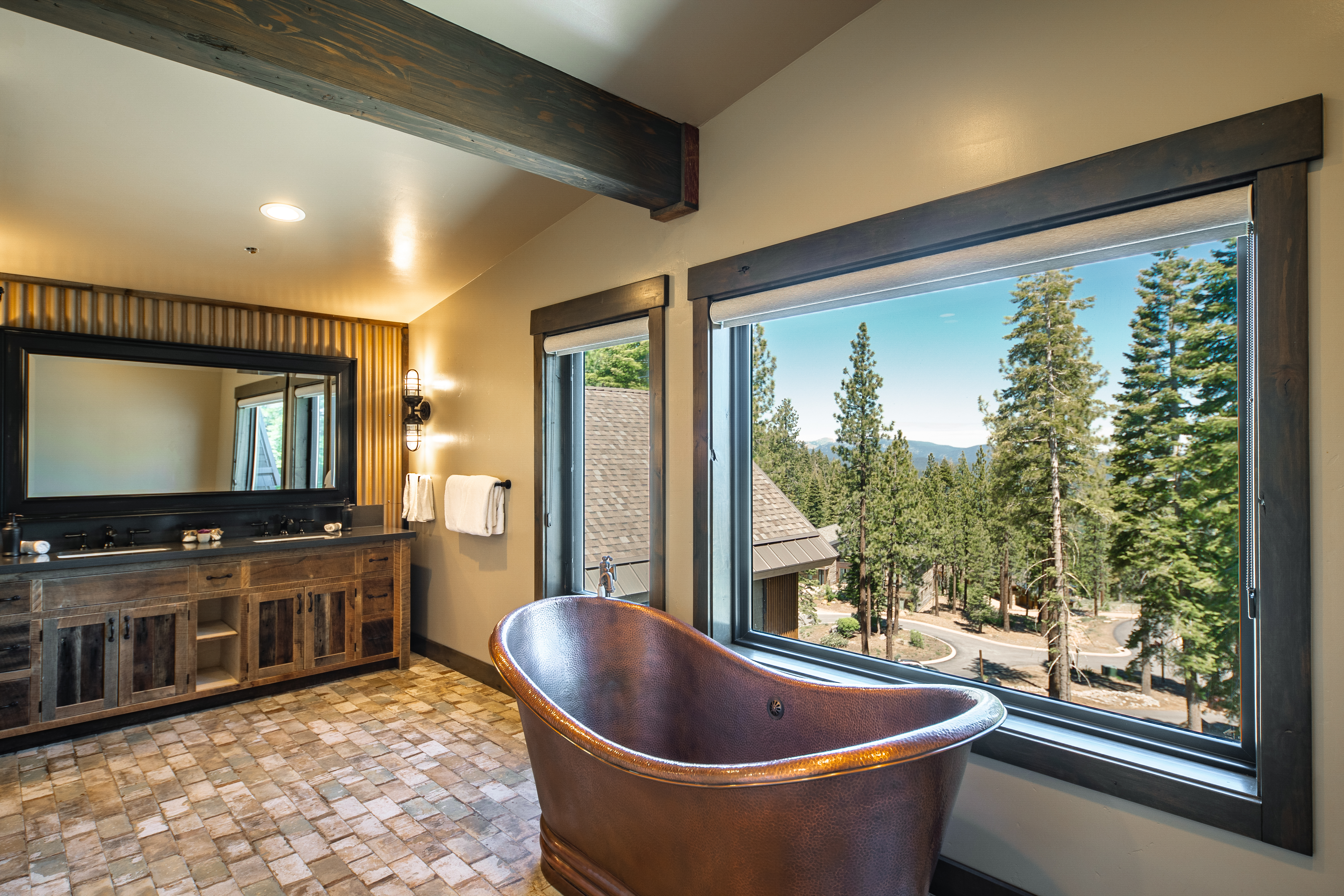 This is a luxurious primary bathroom featuring a copper freestanding bathtub positioned near a large picture window with views of a wooded landscape. The bathroom also includes a rustic wooden vanity with dual sinks, complementing the overall natural and upscale aesthetic. The contrasting textures and the serene outdoor view create a relaxing and spa-like ambiance.