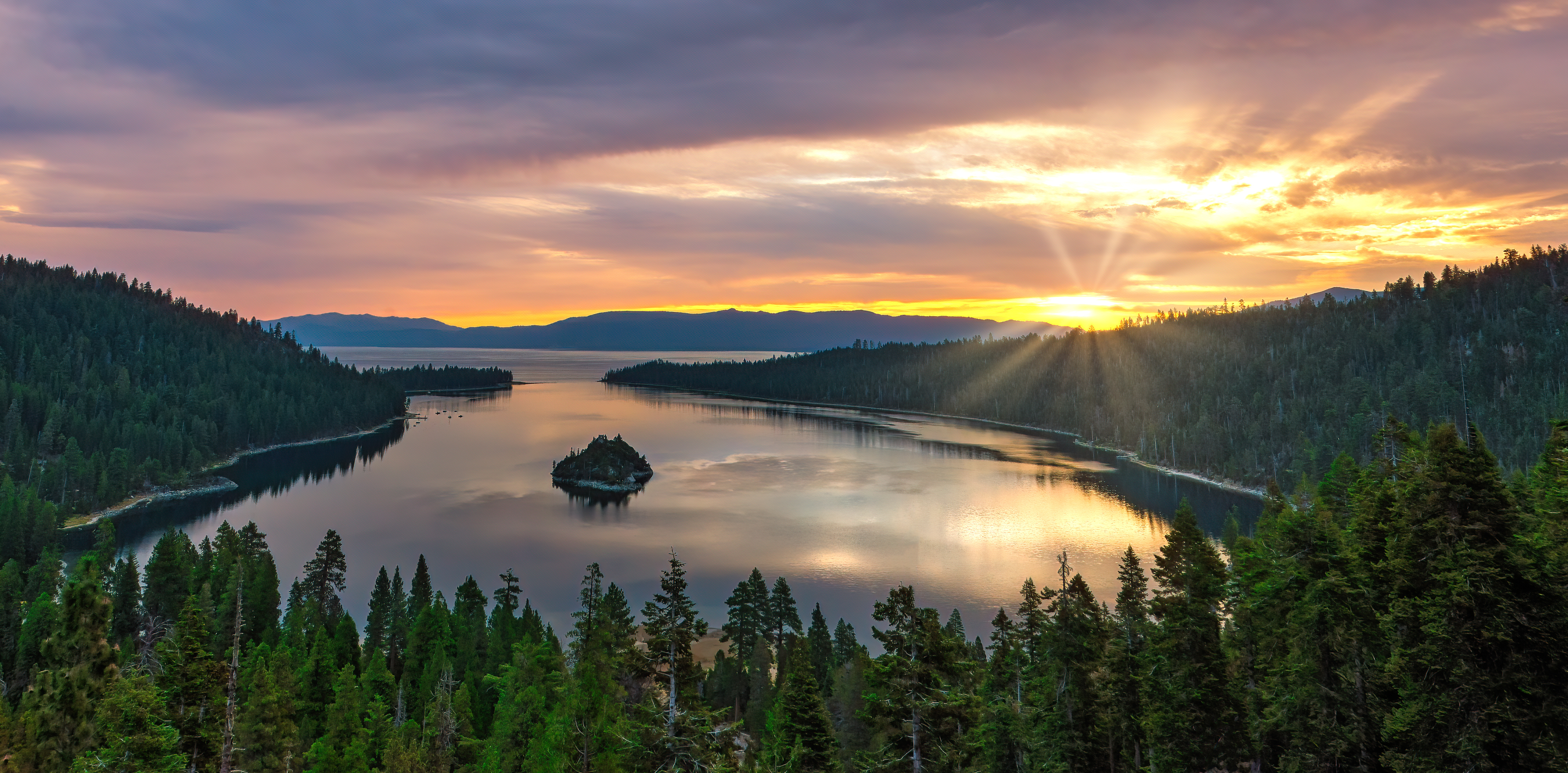 This aerial view captures a serene lake surrounded by lush, forested hillsides at sunset, creating a picturesque and tranquil setting. A small island sits in the lake's center, contributing to the landscape's scenic appeal. The warm, golden light reflecting on the water enhances the overall beauty and feeling of peacefulness, likely appealing to buyers seeking a retreat or nature-oriented property.