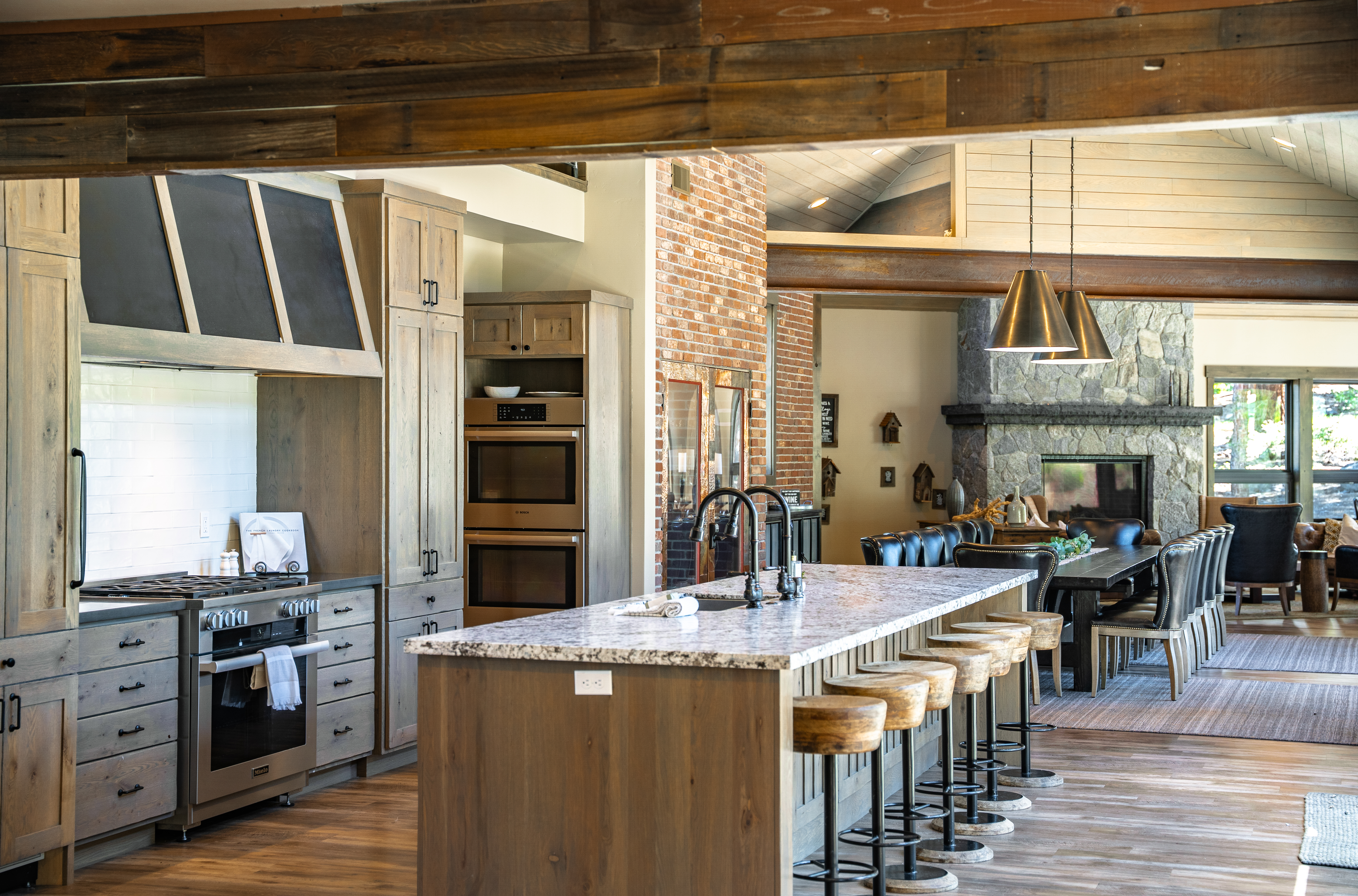 This is a spacious, well-appointed kitchen with rustic contemporary design elements. Notable features include the large center island with bar stool seating, custom cabinetry, and stainless steel appliances, including double ovens and a modern range. The kitchen flows openly into the dining and living areas, creating an inviting and cohesive living space.
