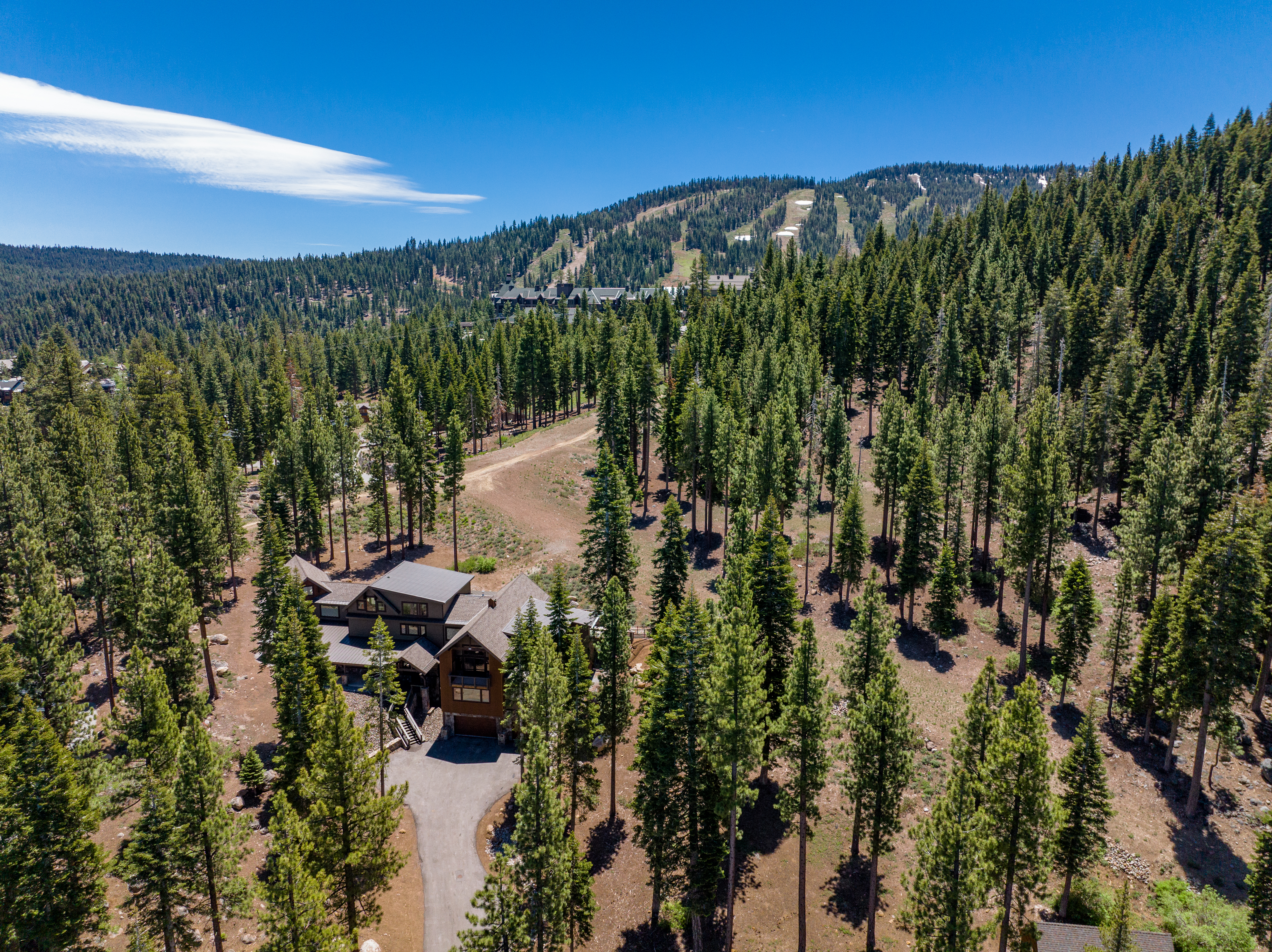 This aerial view showcases a luxurious mountain home nestled among tall pine trees. The property features a spacious driveway and a well-maintained exterior. In the background, ski slopes can be seen, offering potential for winter recreation access.