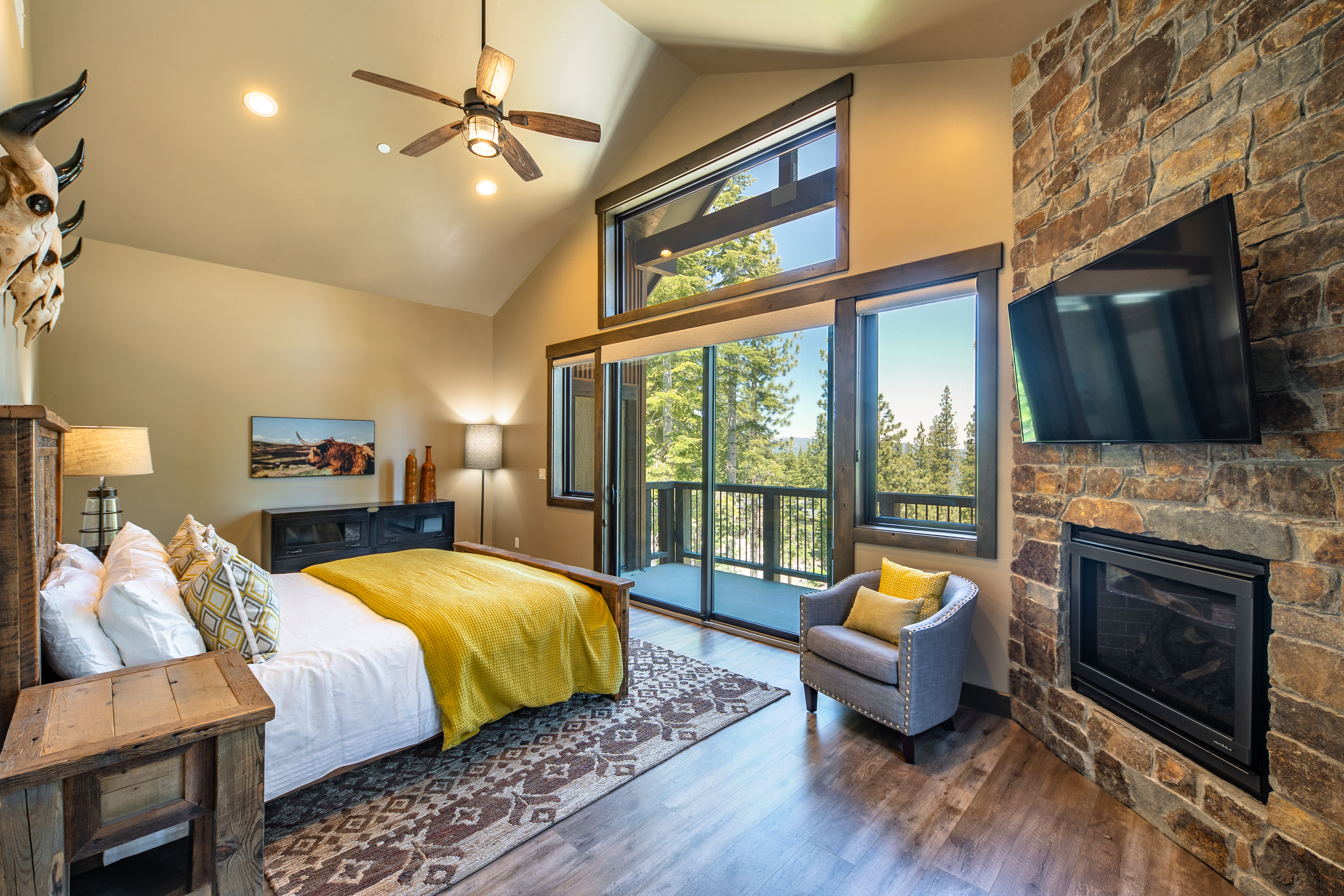 This is a primary bedroom showcasing a rustic yet luxurious style. The room features a bed with a yellow throw blanket, a stone fireplace with a mounted TV, and a large window with a balcony view. The design incorporates natural elements like wood and stone to create a warm and inviting atmosphere.