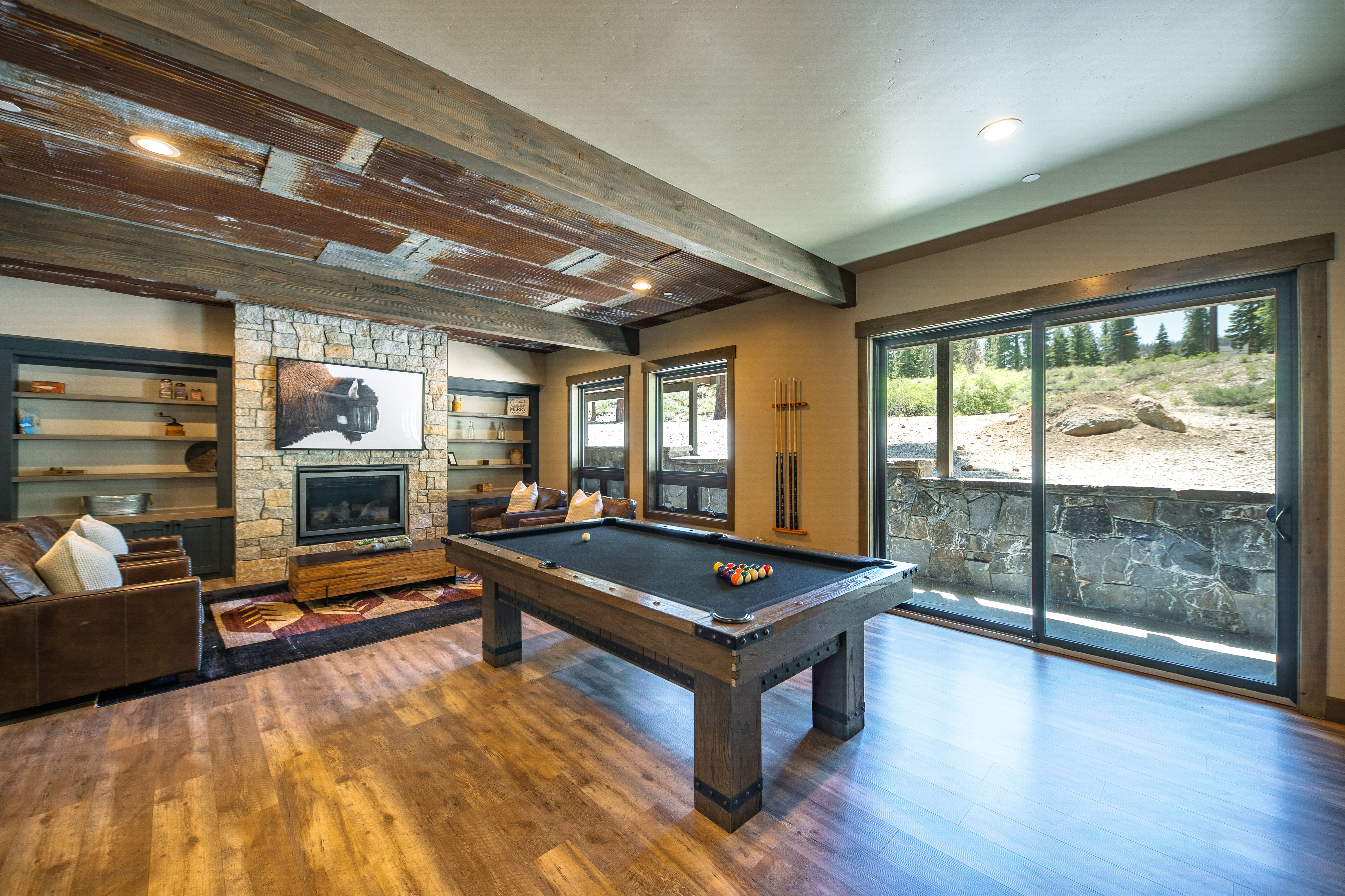 This is an interior shot of a living room featuring a pool table as a central element. The room has a rustic, lodge-like design with a stone fireplace, wooden ceiling beams, and rich wood flooring. Large sliding glass doors provide ample natural light and a view of the outdoor landscape.