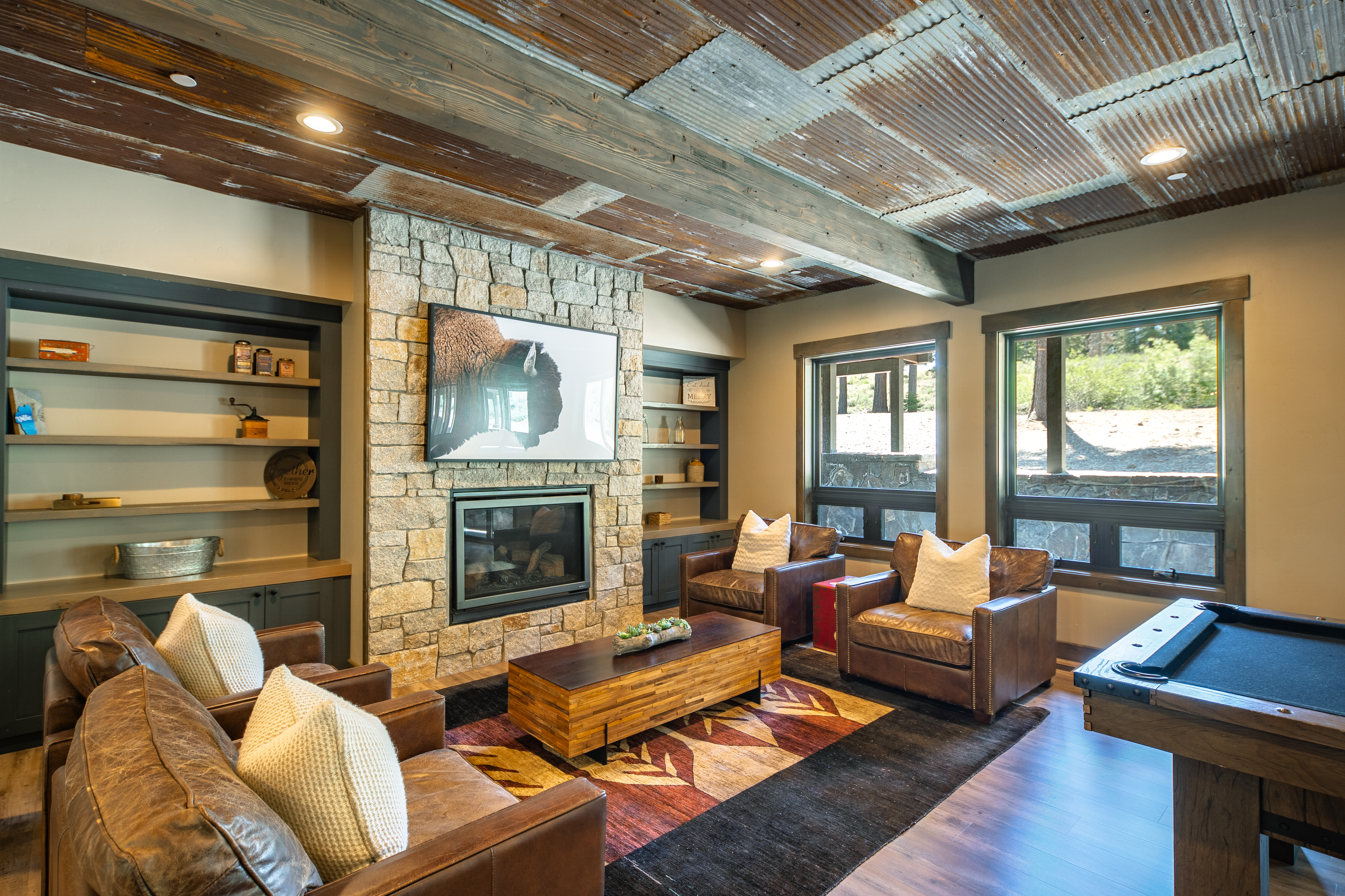 The living room showcases a rustic-chic style with a stone fireplace and built-in shelving units. The room features leather seating, a unique wooden coffee table, and a billiards table, creating an inviting and luxurious space. The ceiling is adorned with corrugated metal accents that add to the room's distinctive charm.