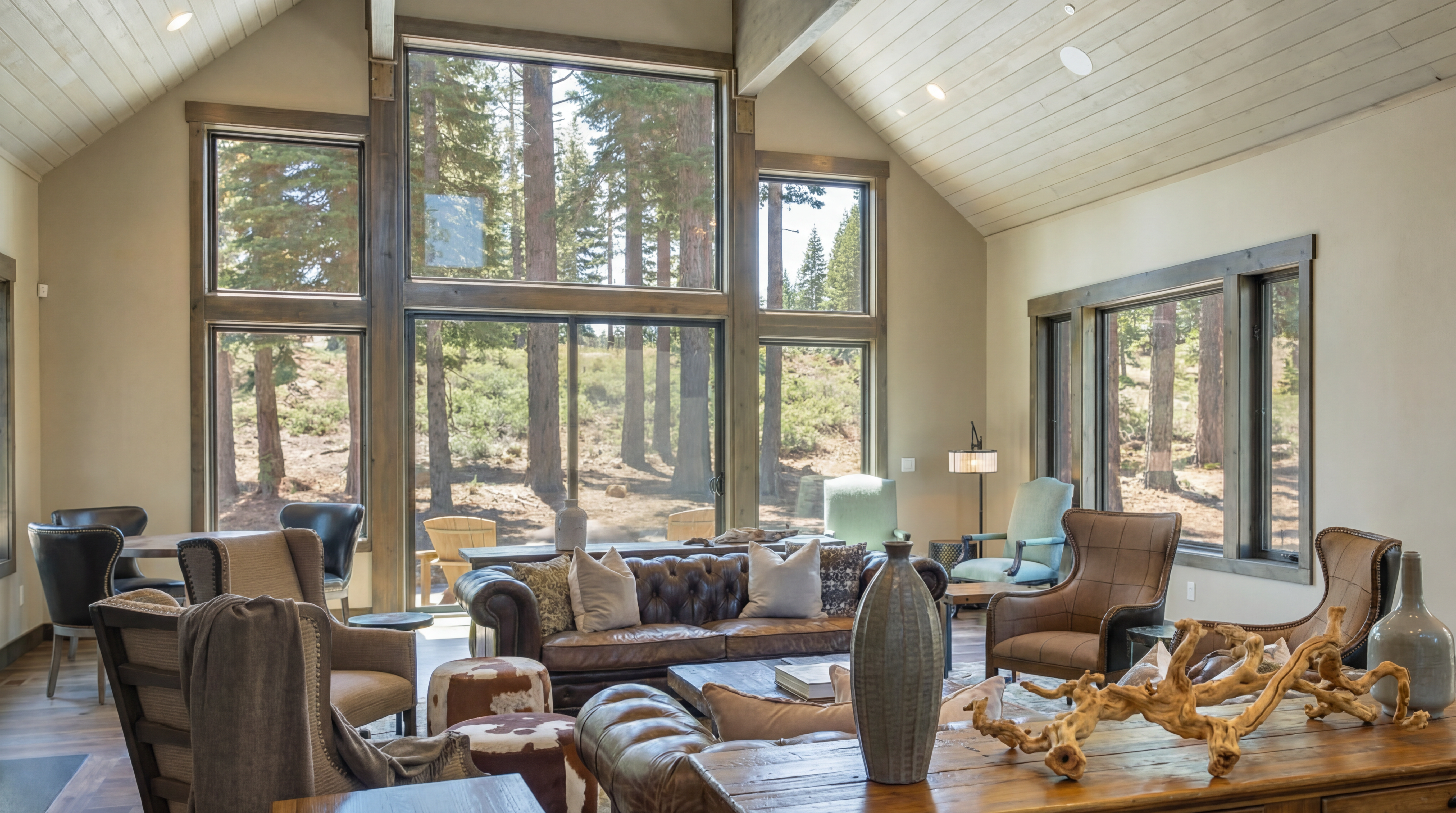 This is an interior shot showcasing a spacious living room with a rustic yet elegant design. Large windows frame a forest view, providing ample natural light. The room features comfortable leather seating, a wooden coffee table with decorative elements, and neutral-toned walls, creating a warm and inviting atmosphere perfect for relaxation and entertaining.