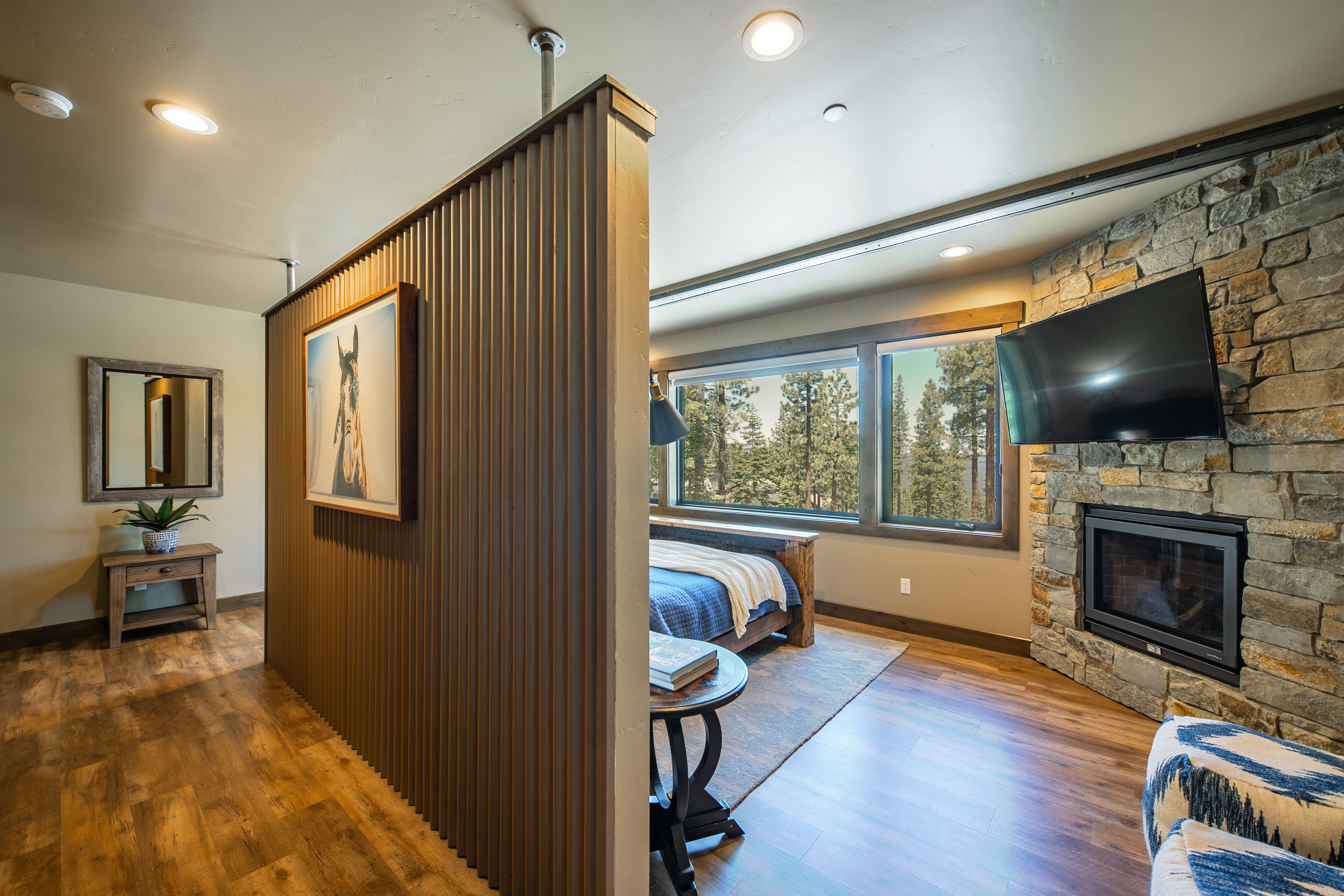 This is a primary bedroom featuring a stone fireplace, flat screen television, and large window with a view of the outdoors. A wooden feature wall partially separates the bedroom from another room. The flooring is hardwood.