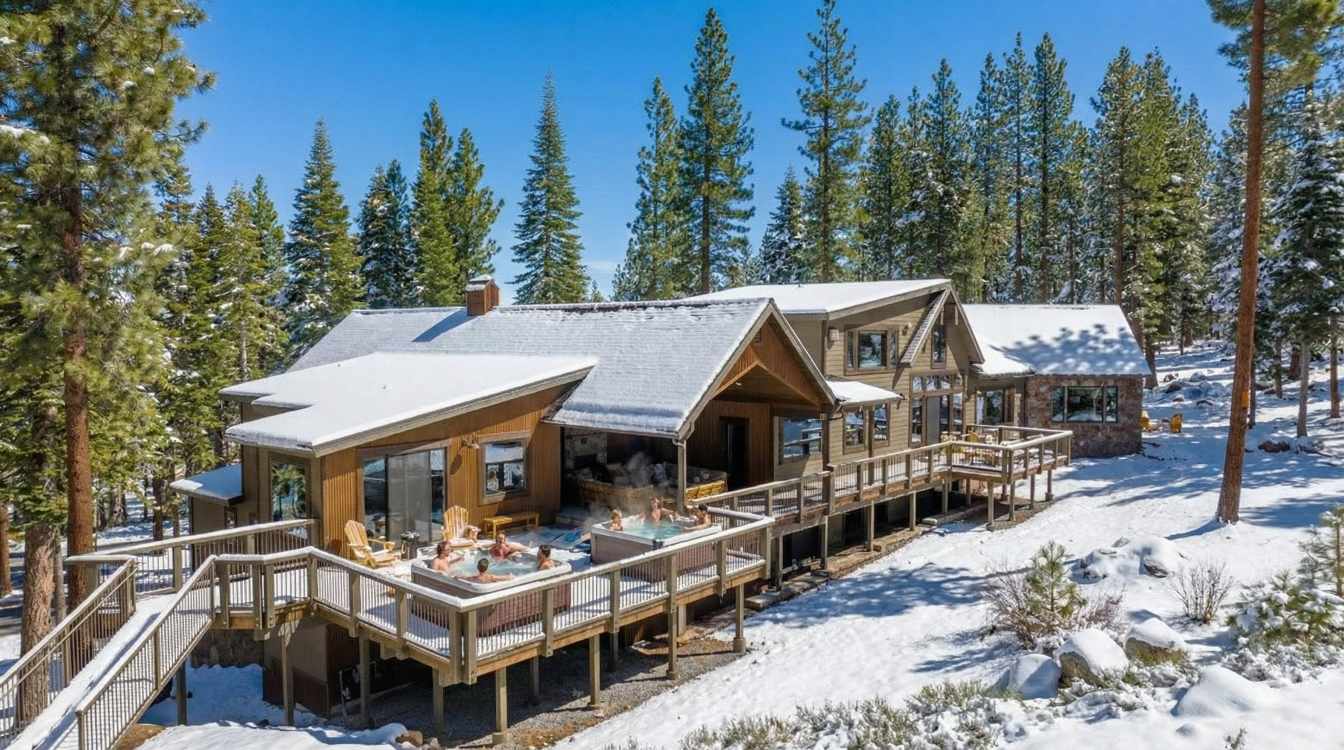 This photograph showcases the rear exterior of a luxurious mountain home, focusing on the expansive deck with hot tubs and scenic views of the surrounding snow-covered evergreen forest. The architecture features wood siding and a roof covered in snow, blending harmoniously with the natural landscape. The presence of people enjoying the hot tubs adds a sense of relaxation and leisure.