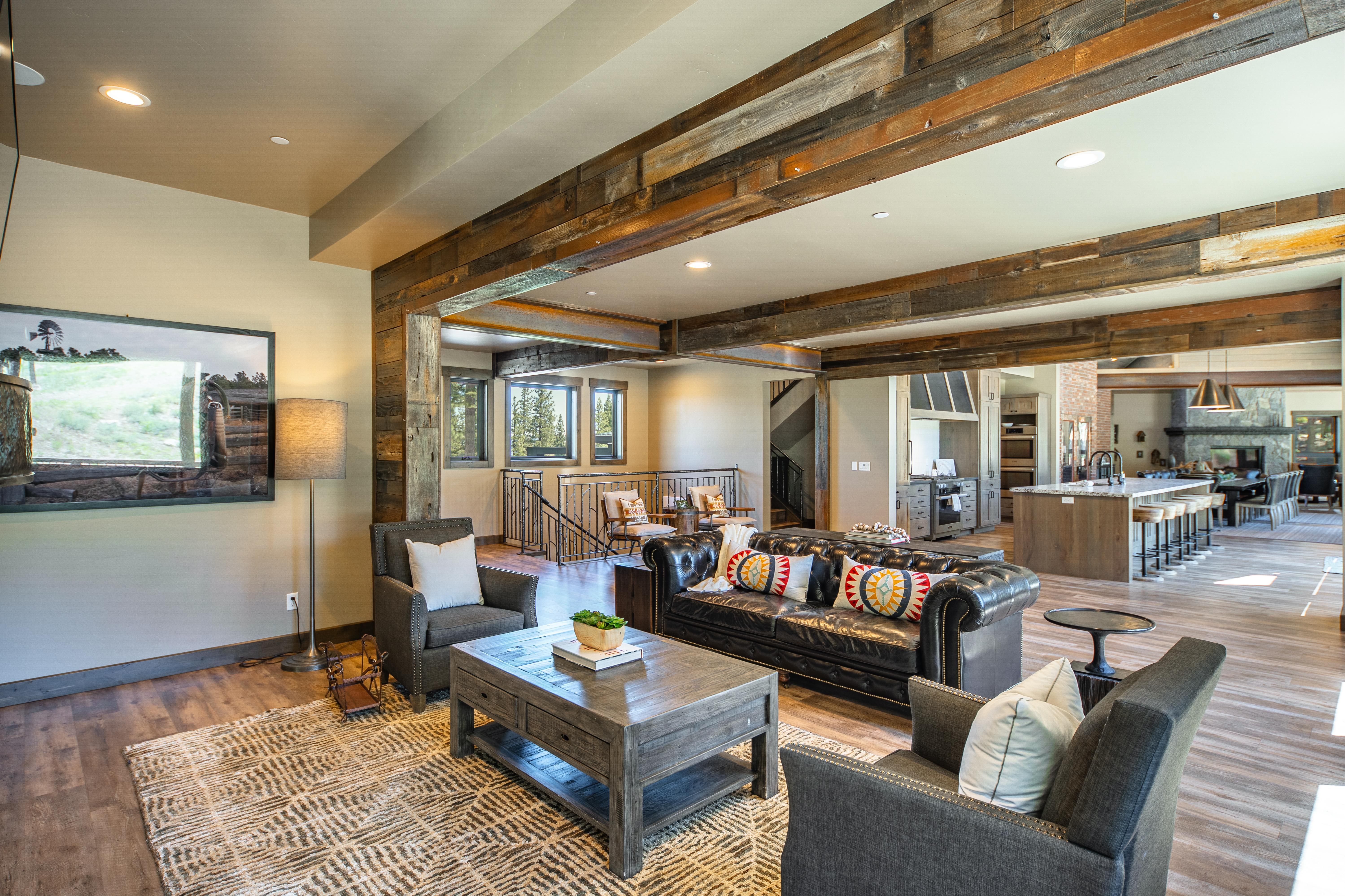 This is a spacious living room with a rustic-contemporary design. It features exposed wooden beams, hardwood floors, and comfortable seating including a leather couch and upholstered armchairs. A decorative rug adds warmth to the space, and the open layout connects seamlessly to the kitchen.