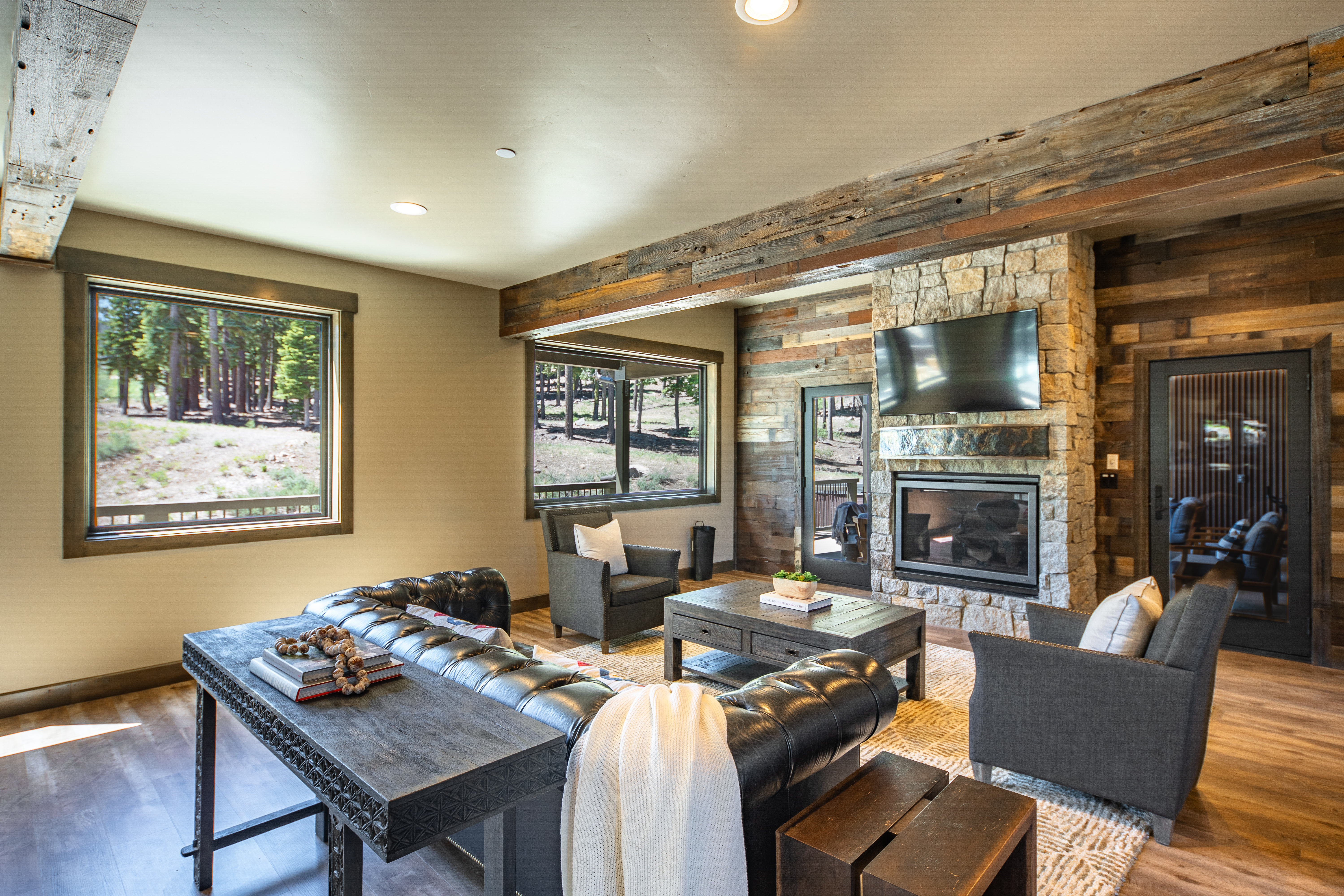 This is an inviting living room featuring a stone fireplace with a mounted TV, wood-paneled walls, and comfortable seating arrangements. The room is filled with natural light from large windows that offer views of the outdoors. The overall impression is one of rustic elegance and warmth.