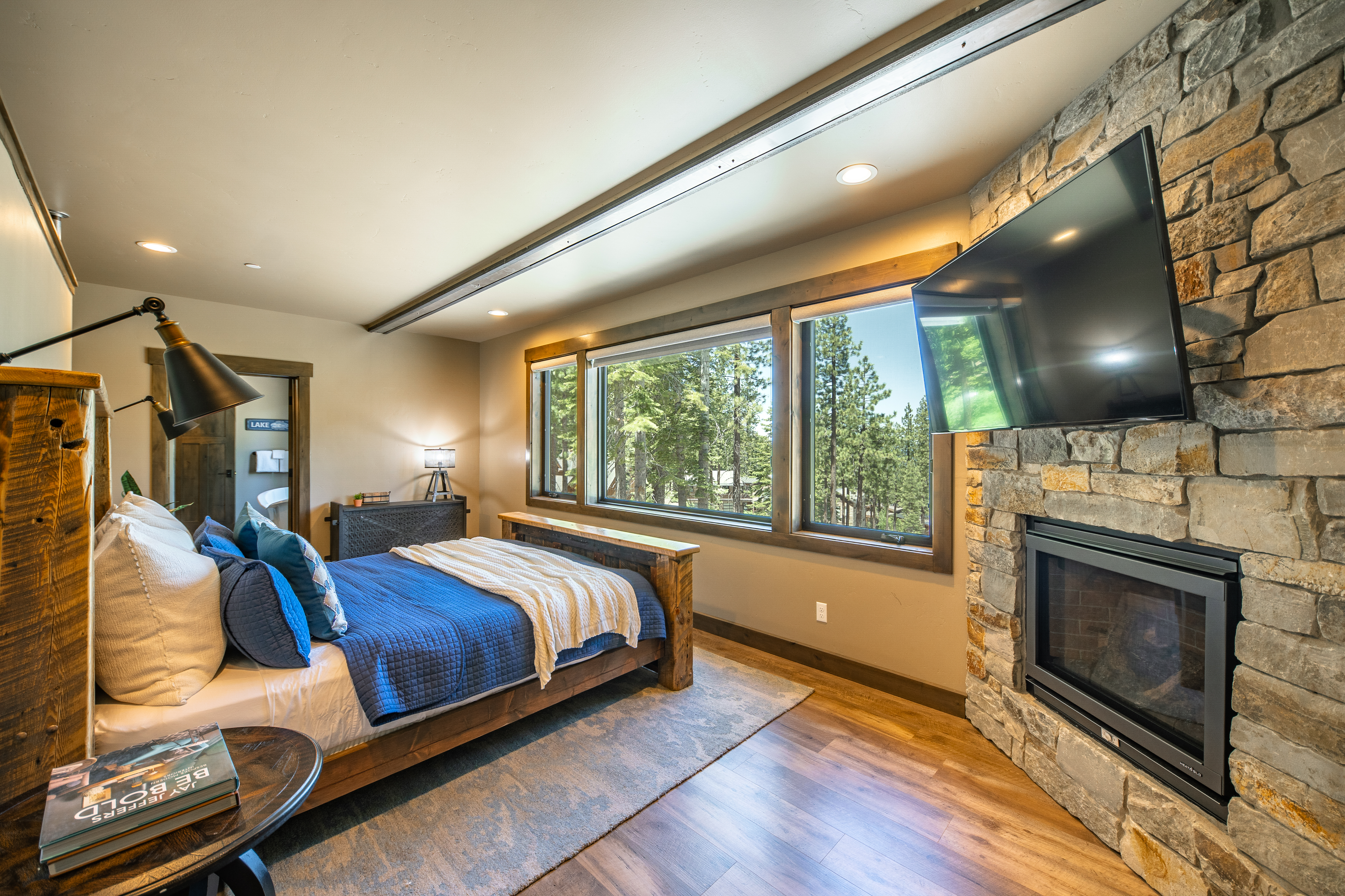 This is a cozy, well-lit primary bedroom featuring a rustic wooden bed with blue bedding, complemented by a stone fireplace with a mounted television. Large windows offer a view of trees, enhancing the room's serene atmosphere. The room features a blend of rustic and modern elements.