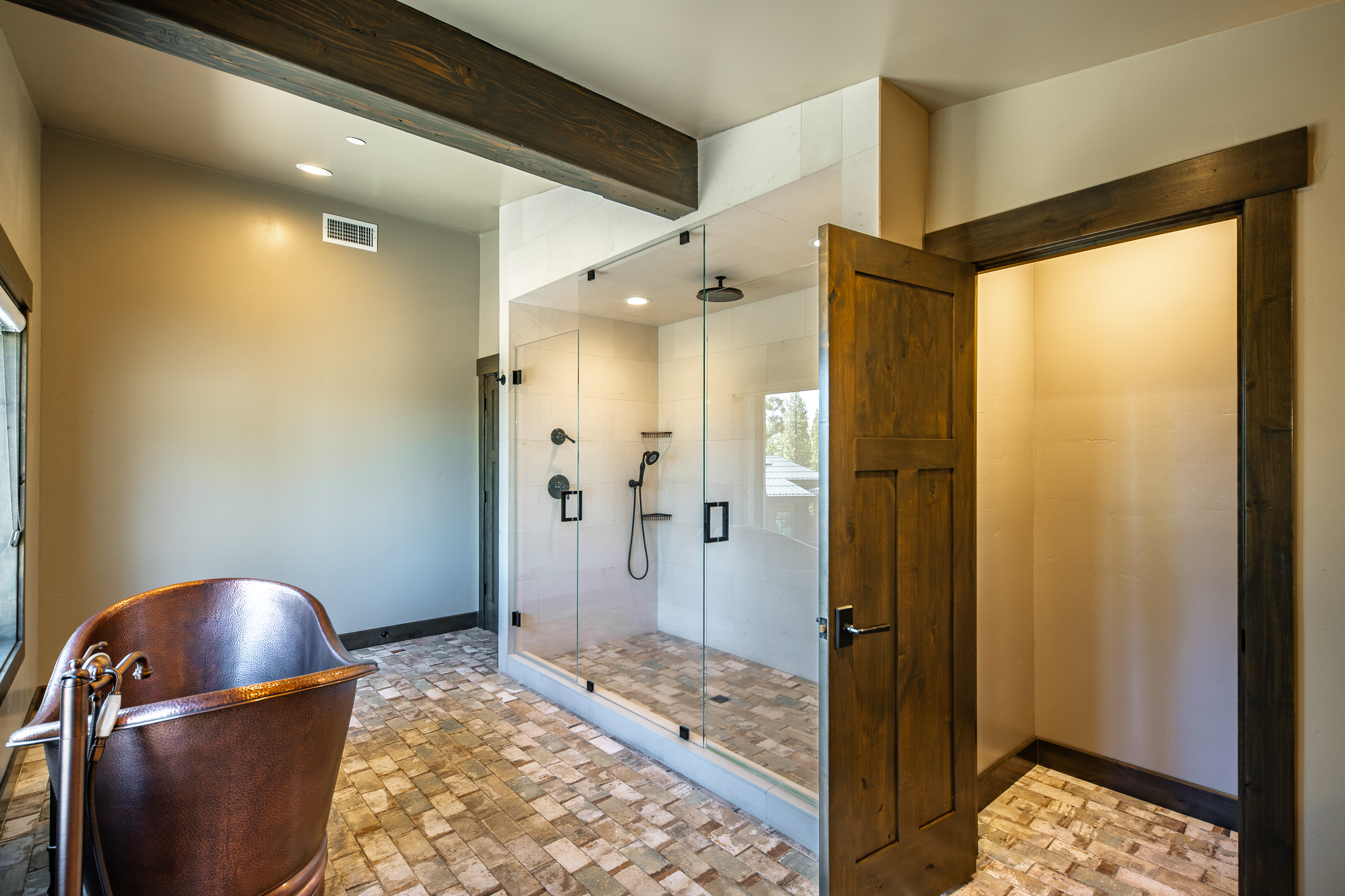 This primary bathroom showcases a luxurious shower with glass enclosure, and a striking copper bathtub next to a window. The space is accented with dark wood beams and door frames, complemented by unique tile flooring. The overall impression is one of rustic elegance and comfort.