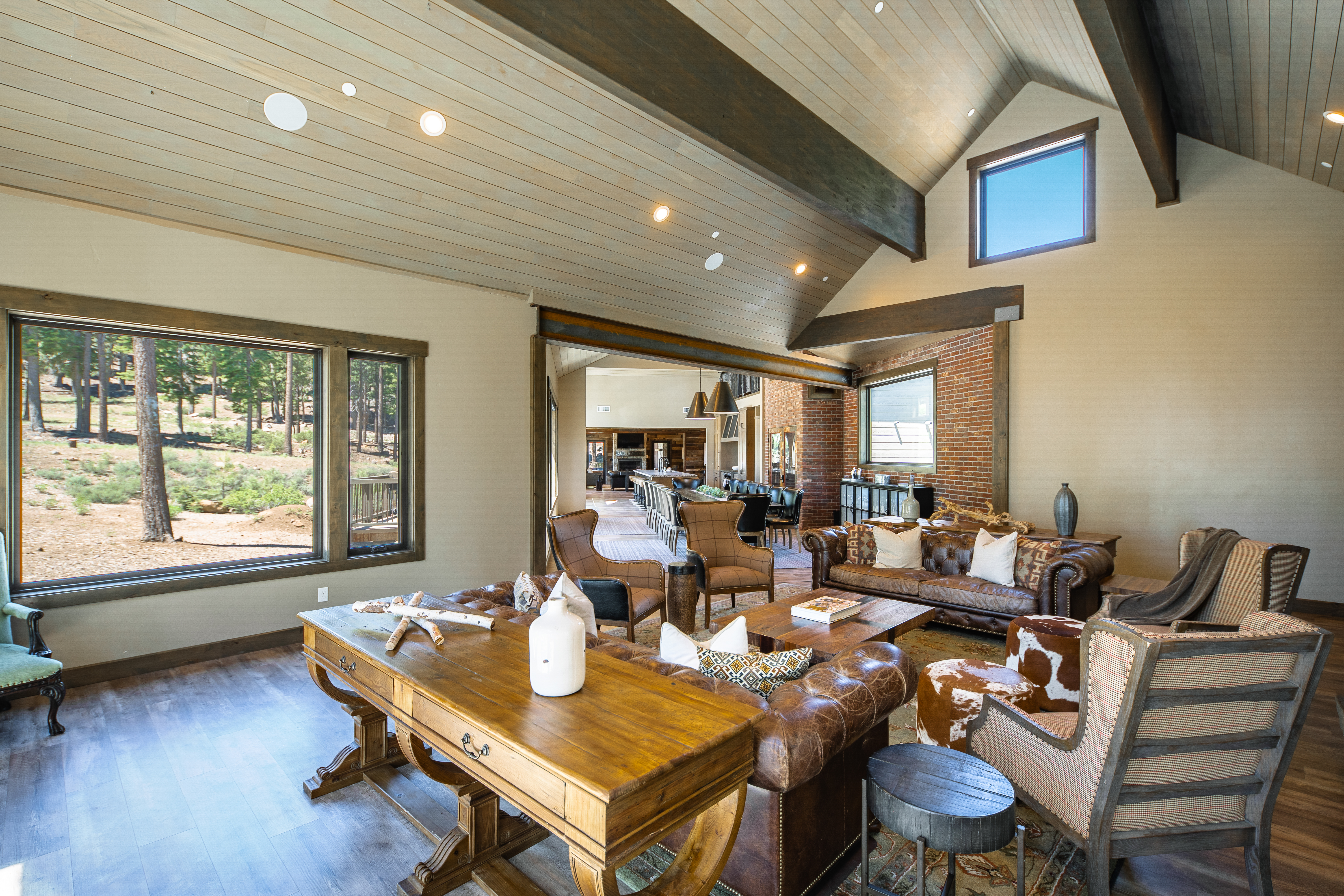 This is a spacious living room with a rustic yet elegant style. It showcases hardwood floors, exposed beams, and a large window overlooking the outdoors. The room is furnished with leather sofas, armchairs, and a wooden desk, creating a warm and inviting atmosphere perfect for relaxation and entertaining.
