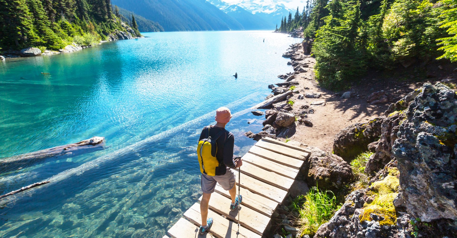 A scenic landscape featuring a hiker crossing a wooden bridge over clear turquoise lake, potentially indicating access to scenic trials from a property. The natural beauty of the location, with the lush forest and snow-capped mountains in the background, could be a major selling point.
