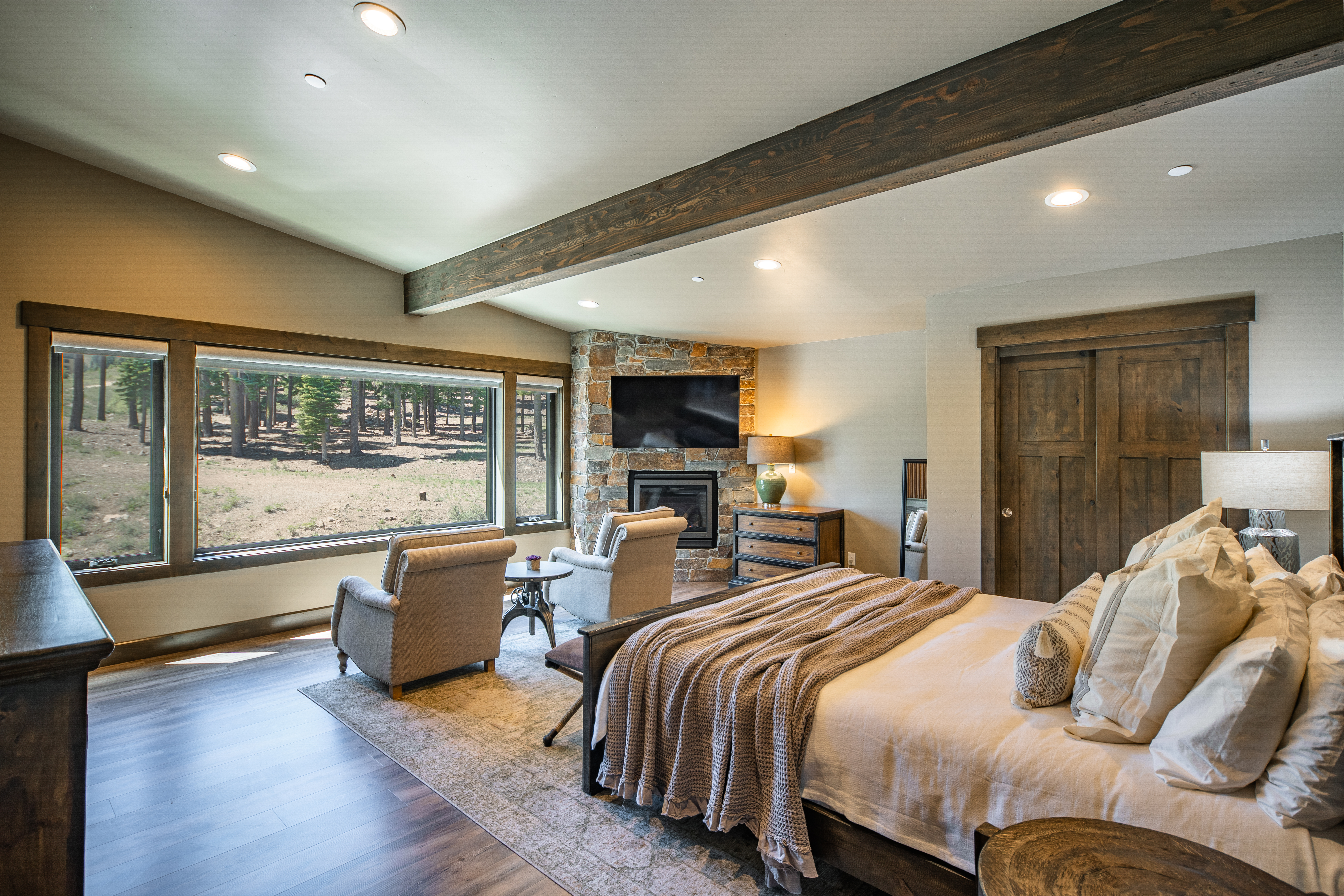 This is a master bedroom featuring a stone fireplace with a television mounted above, two cozy armchairs, and a large window with views of the outdoors. The room has a rustic yet modern aesthetic with wooden beams, trim, and a comfortable looking bed. Natural light floods the space, creating a warm and inviting atmosphere.