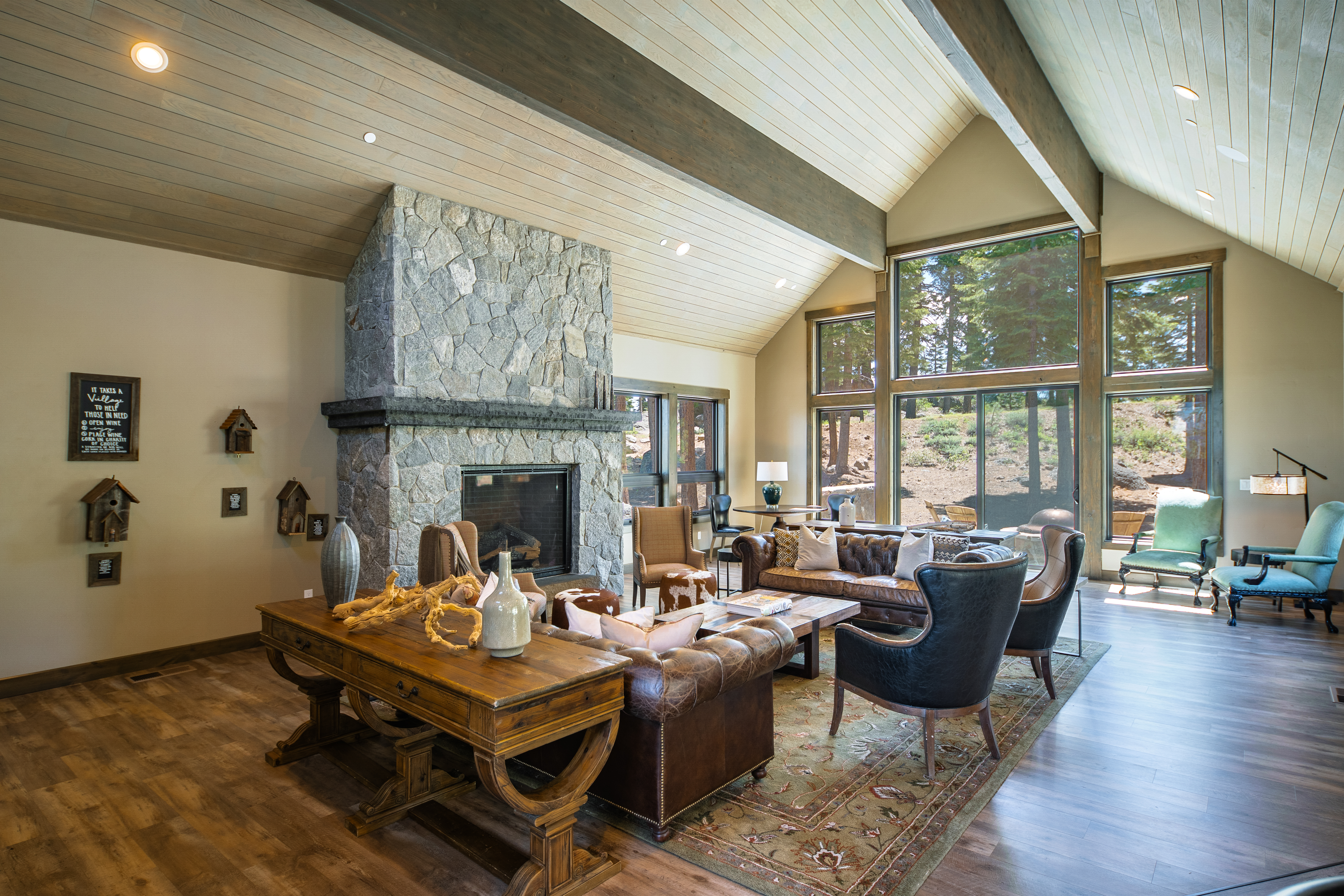 This is a shot of a traditional-style living room with a stone fireplace, wooden beams, and hardwood floors. The room features leather sofas and armchairs arranged around a coffee table layered on a patterned rug, indicating a comfortable and inviting gathering space. Large windows allow for views of the natural scenery, adding to the room's charm.