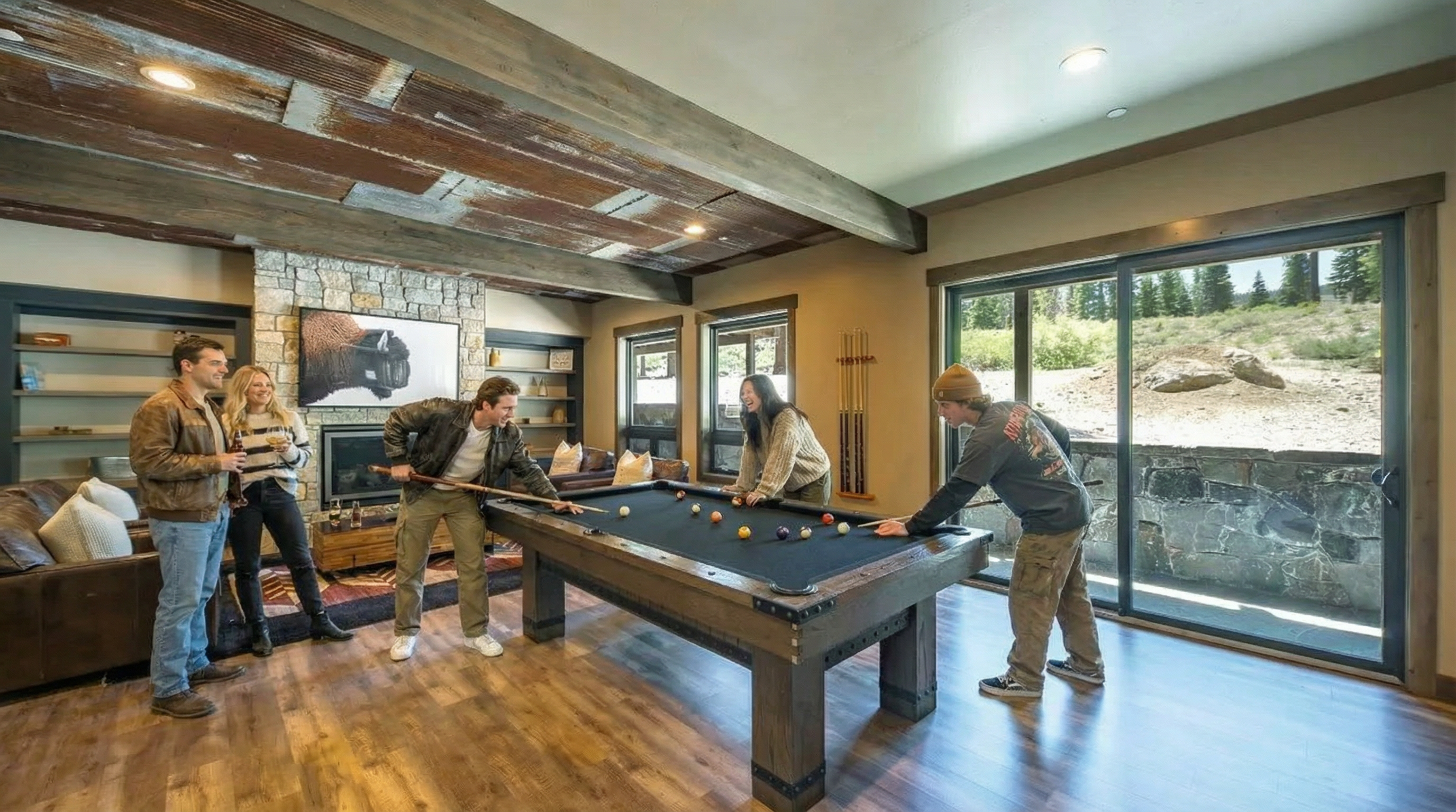 This interior showcases a well-lit living room with a rustic-modern aesthetic, featuring a pool table, stone fireplace, and wood beam ceiling. The open layout, combined with natural light streaming in from large windows and a sliding glass door, creates an inviting space for recreation and relaxation. Hardwood flooring and neutral wall colors enhance the room's warmth and comfort.
