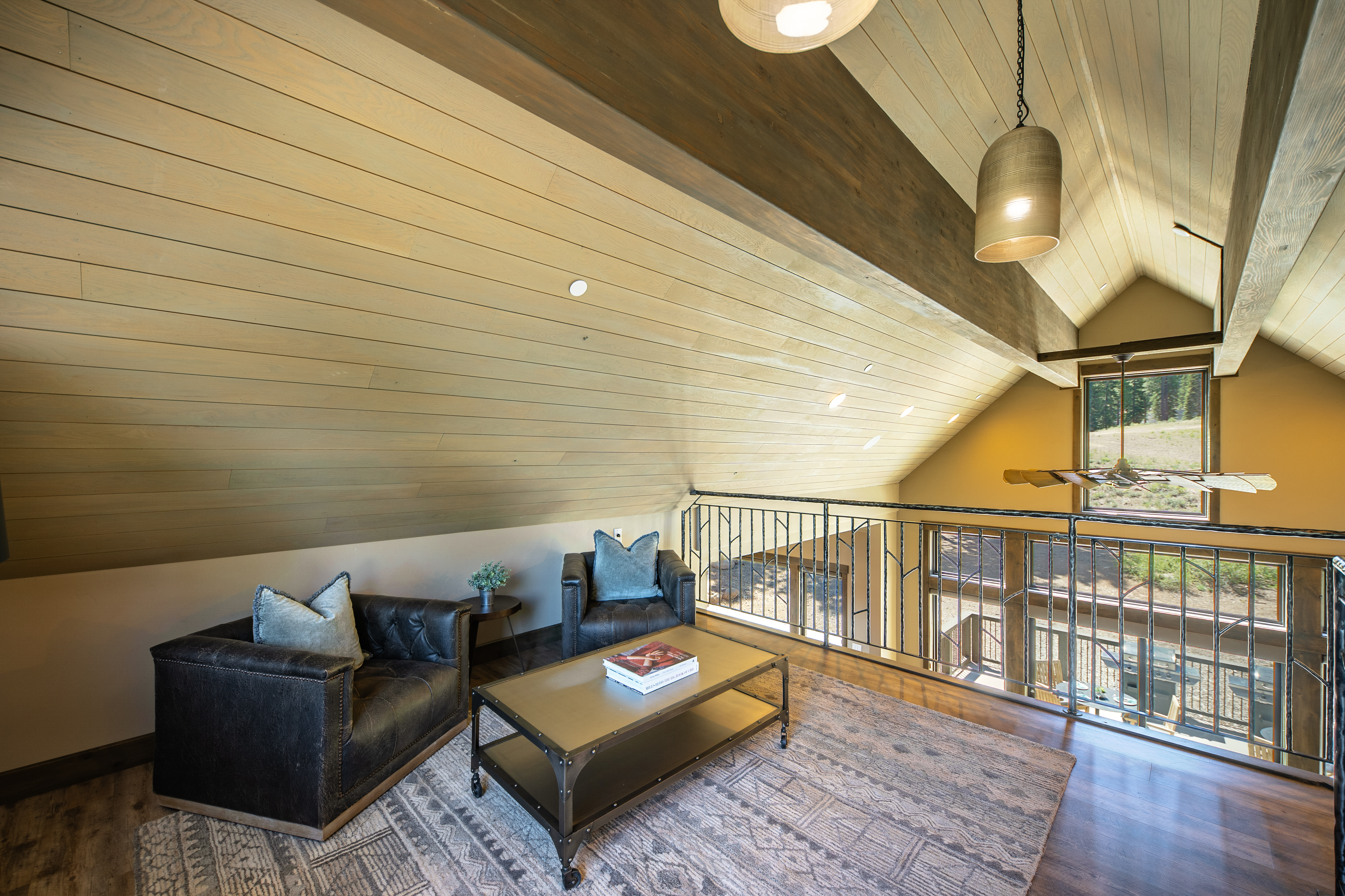 This is a cozy living room area in an attic space with wood paneled walls and ceiling. Two leather armchairs with plush pillows, a wooden coffee table with books, and an area rug create an inviting setting. The room features railing overlooking the floor below and a window that brings in natural light.