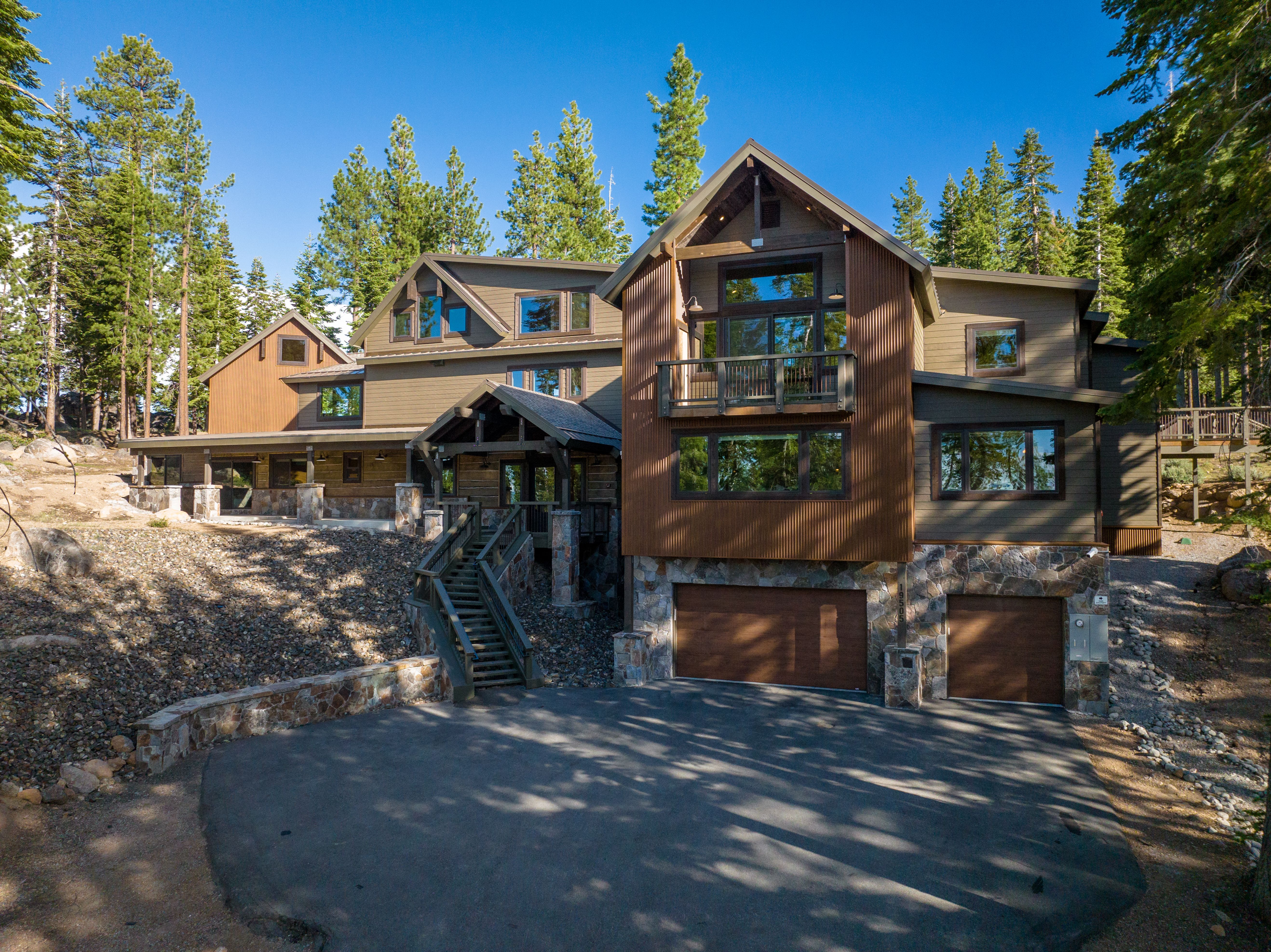 This is a view of a luxury home's facade. The exterior features a blend of wood and stone, lending a rustic yet modern feel. A well-maintained driveway leads to a two-car garage, and there are stairs leading up to what appears to be a spacious entryway.