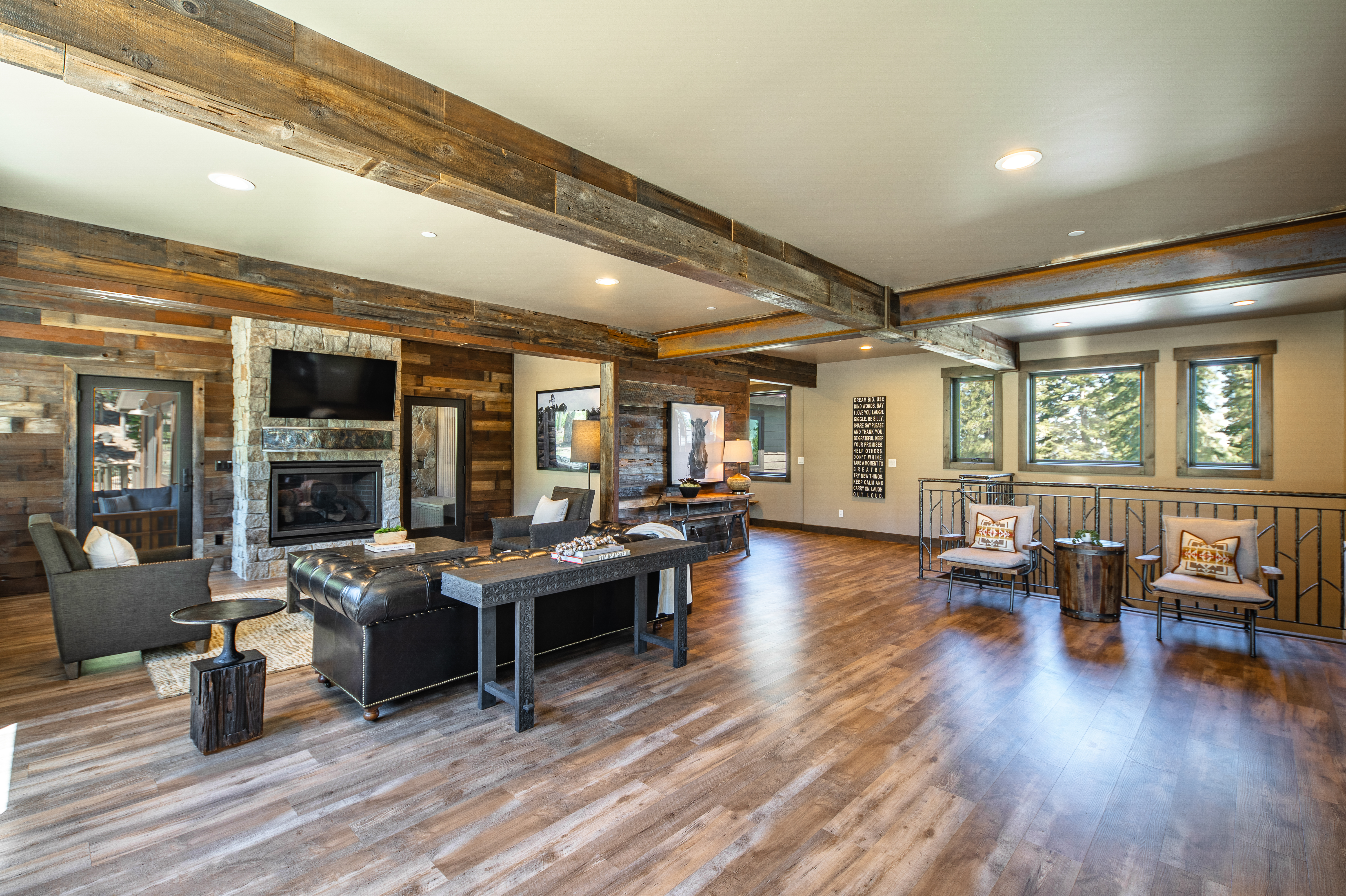 This is a spacious living room area. The room features hardwood floors, exposed wooden beams across the ceiling, and a stone fireplace with a mounted television. There is a comfortable seating area, a console table, and an elevated loft-like space with additional seating and views from the windows.