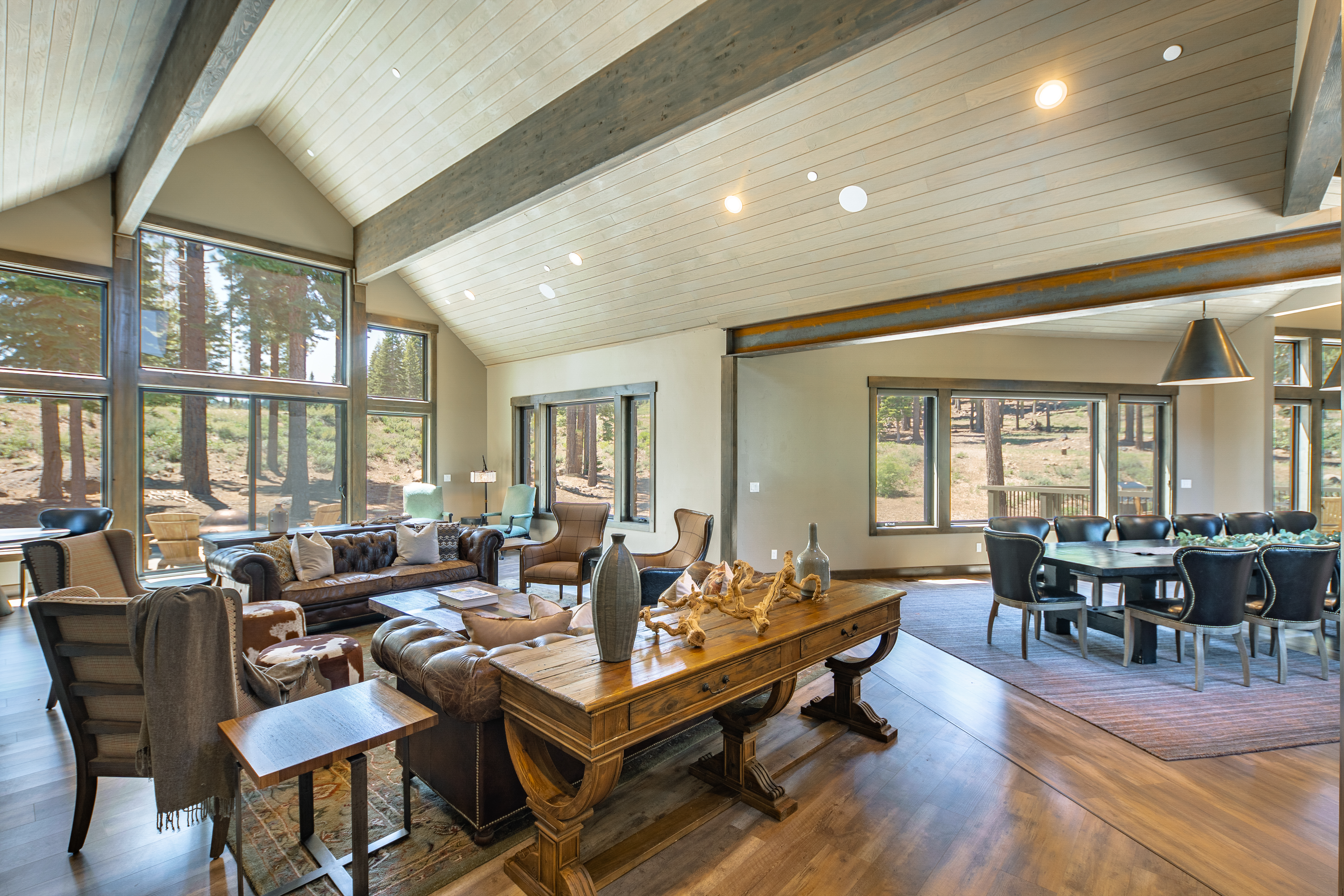 This is a spacious living area featuring a high vaulted ceiling with wooden beams and large windows offering natural light and scenic views. The room is furnished with comfortable leather sofas, chairs, and a wooden console table. An adjacent dining area with a large table and seating is also visible, suggesting an open floor plan, and the overall impression is warm and inviting.