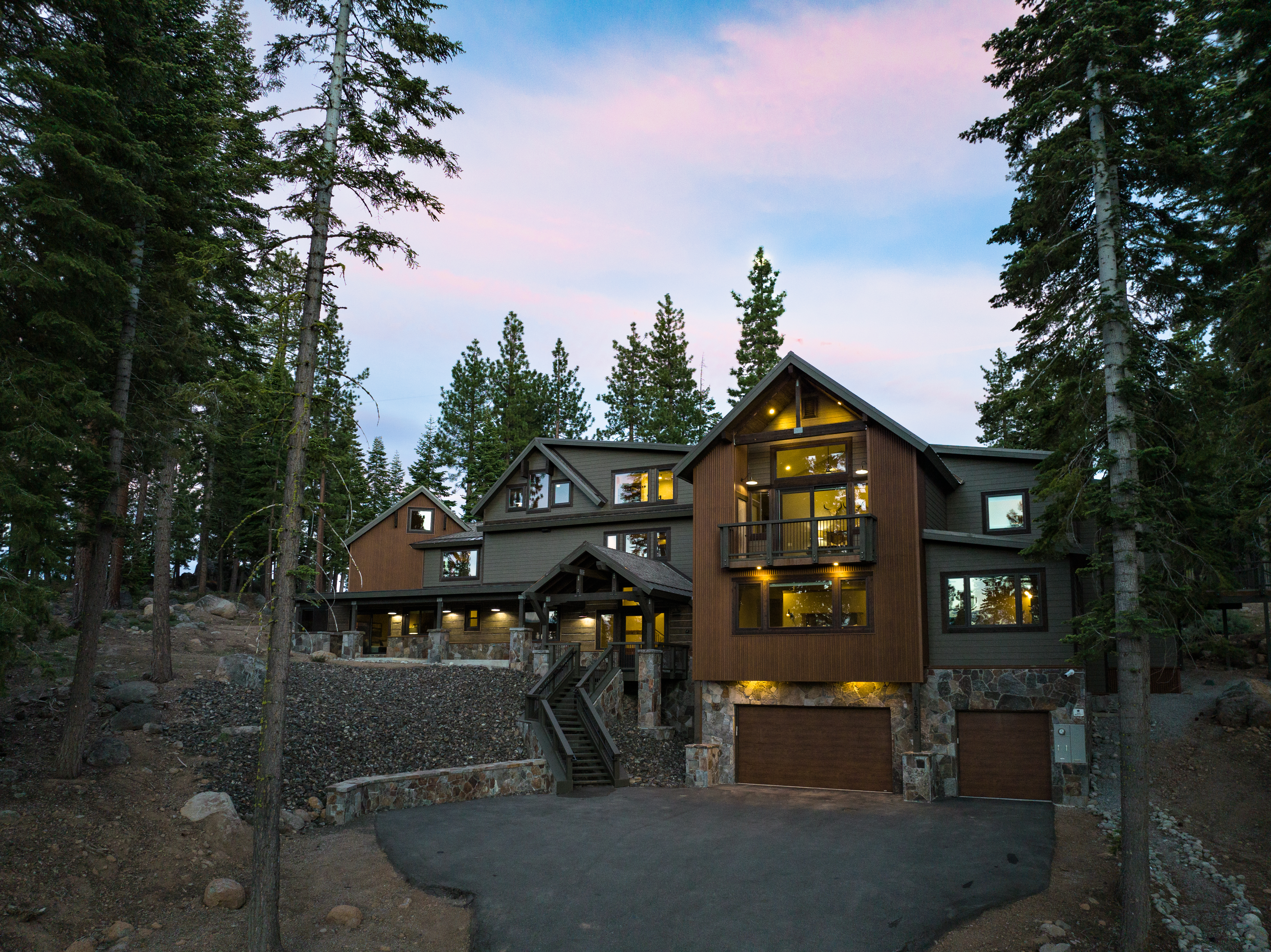The image showcases the front exterior of a multi-level home nestled in a wooded area. The home features a combination of wood siding and stone accents, with a visible two-car garage and multiple windows. A set of stairs leads to the entrance, hinting at a well-maintained property with a touch of rustic charm.