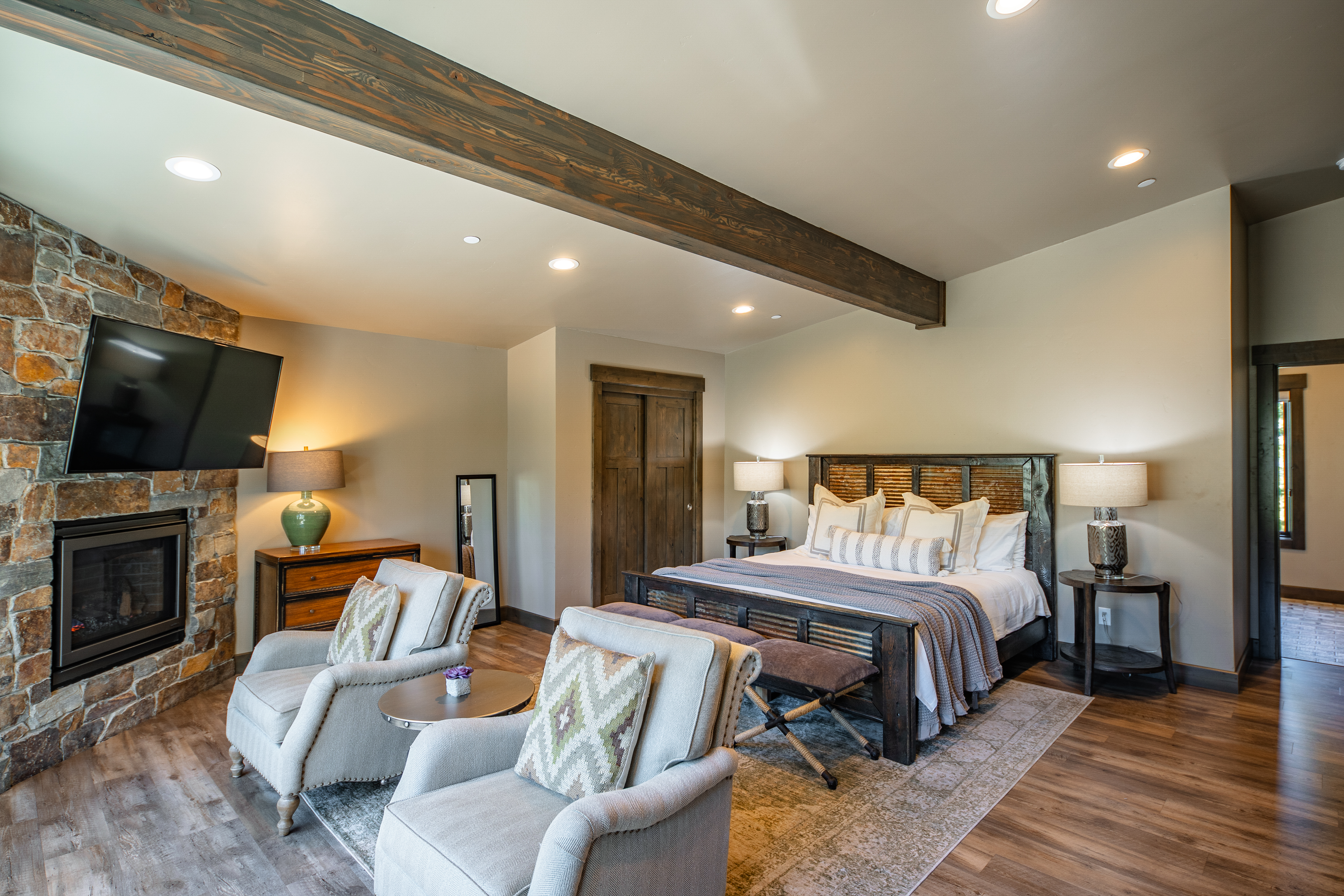 This is a spacious primary bedroom featuring a stone fireplace with a mounted TV, two comfortable armchairs, and a king-size bed with a decorative headboard. The room is decorated in neutral tones with pops of color in the pillows and lamps, creating a cozy and inviting atmosphere. A wooden beam adds a rustic element to this relaxing space.