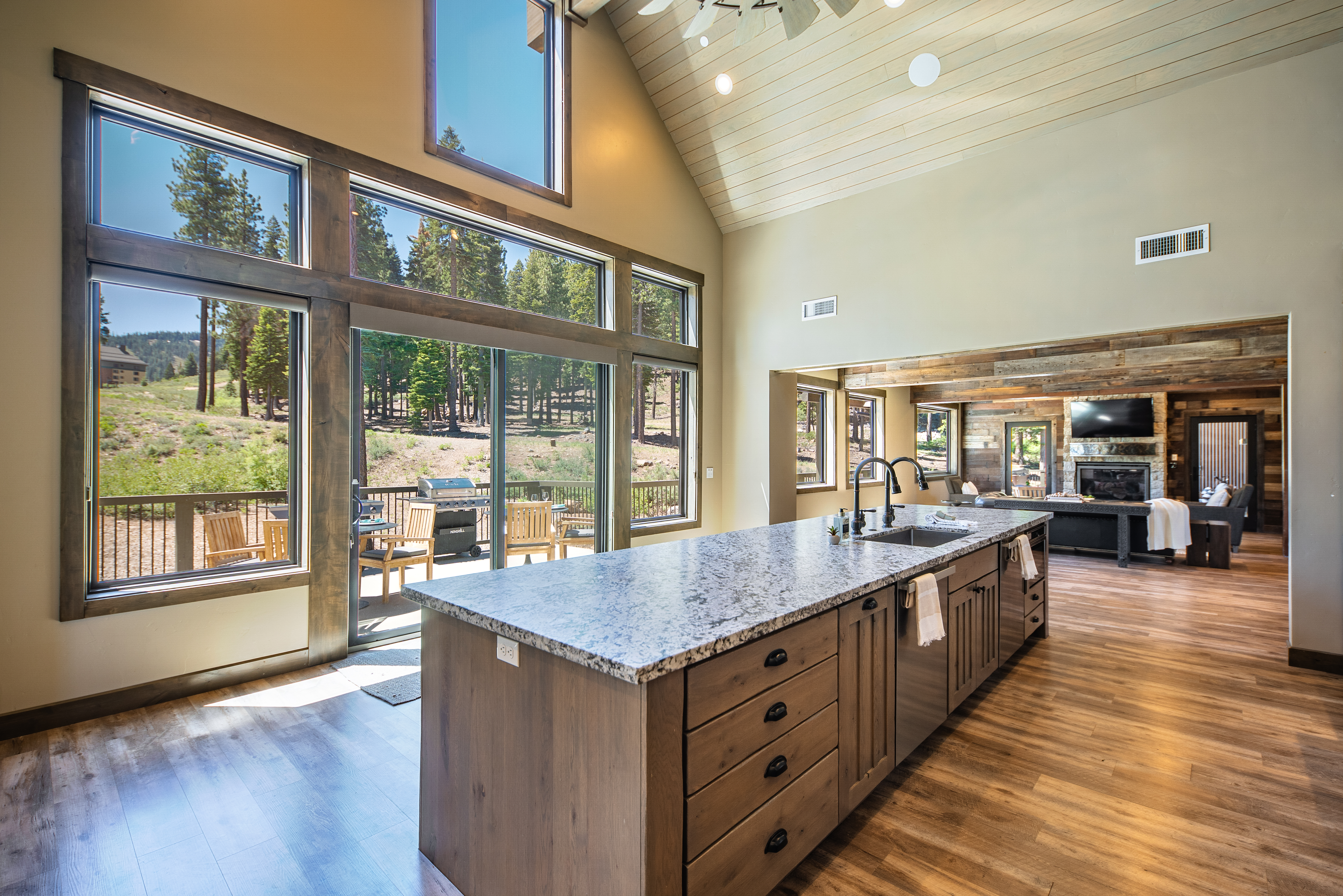This image showcases a well-lit kitchen with a spacious island featuring a granite countertop and dark wood cabinetry. Large windows provide ample natural light and a view of the outdoor scenery. The kitchen seamlessly flows into the living area with wood paneling adding a rustic touch, creating an inviting open-concept living space perfect for entertaining.