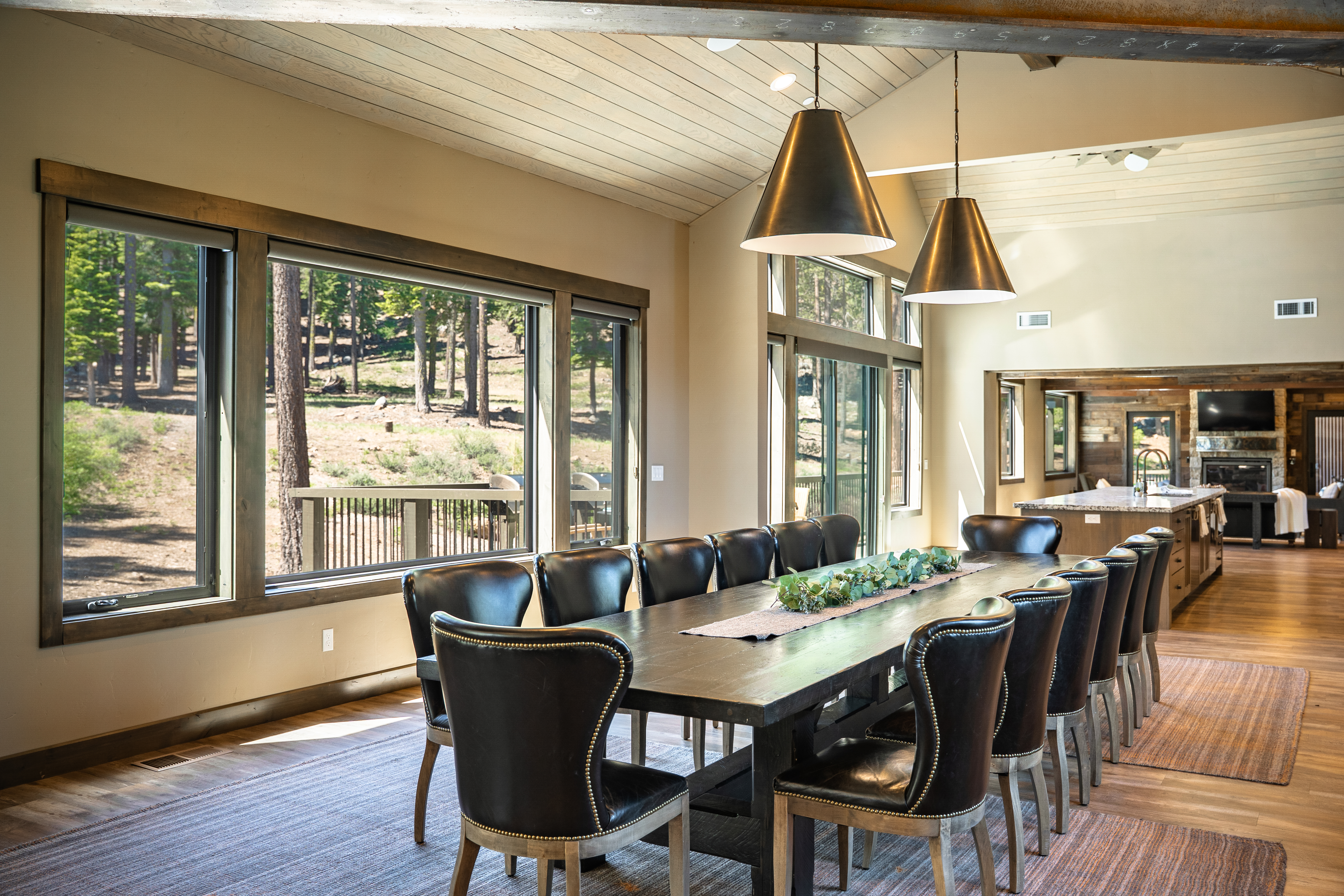 The image showcases a dining room featuring a long, dark wood dining table with seating for several people. Large windows provide natural light and views of the outdoors along with two pendant lights providing additional lighting. The space is decorated with a runner and greenery, and complements a warm and inviting atmosphere.
